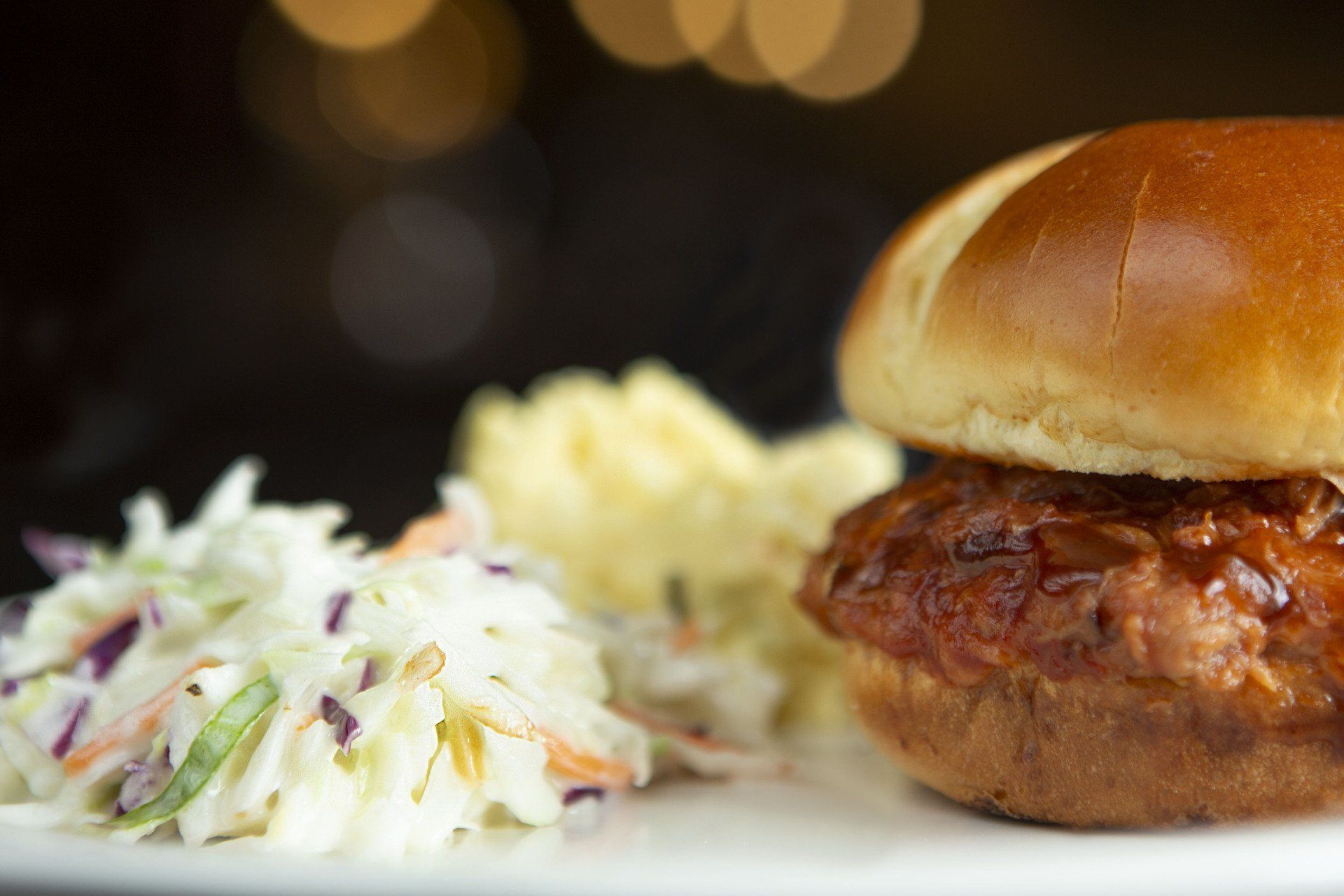 Plated burger with coleslaw and mashed potatoes on a white plate, softly lit background