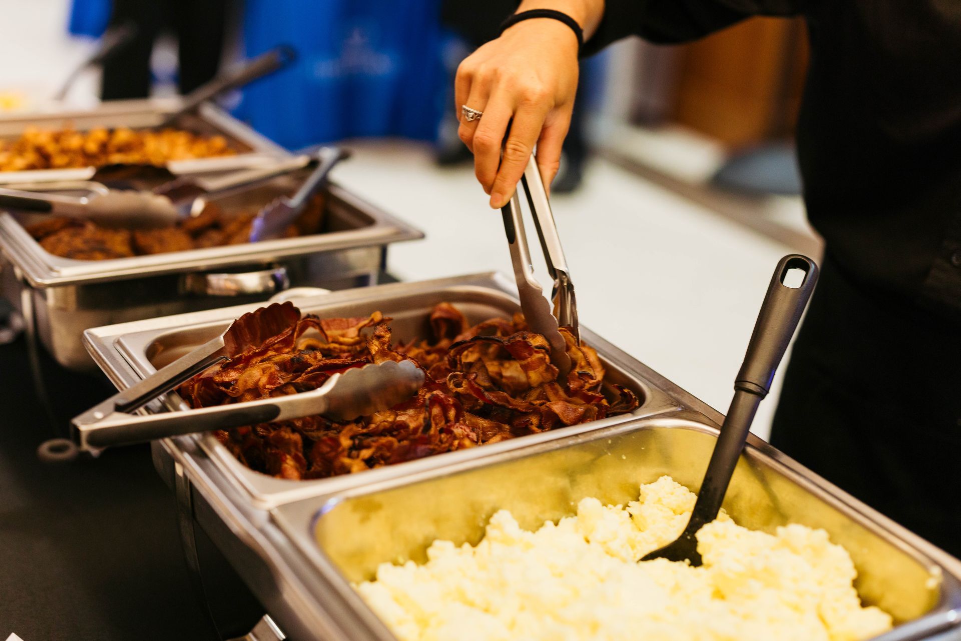 Buffet trays with shredded meat and mashed potatoes; a hand serves food with tongs