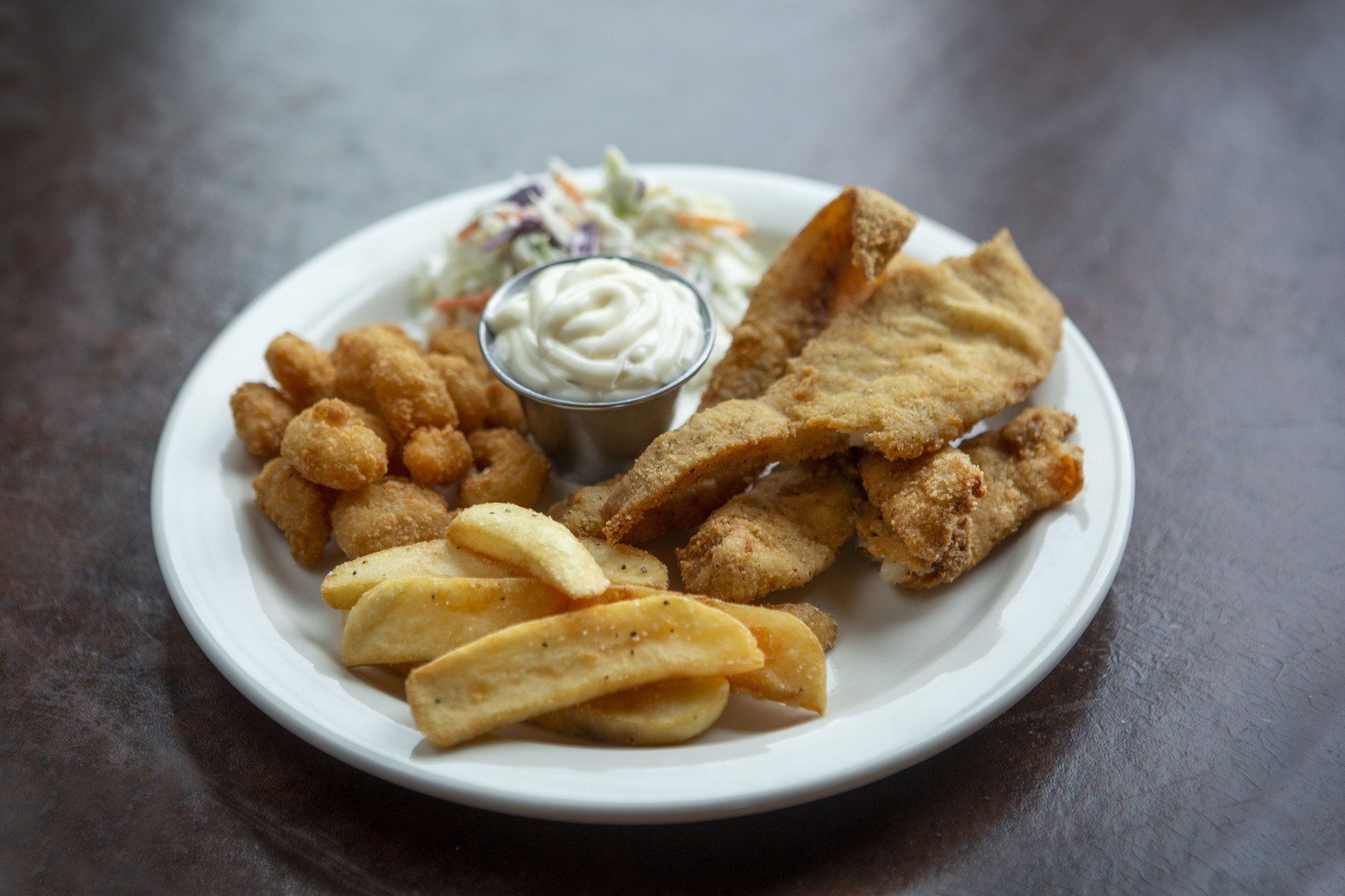 Plate of fried fish, chips, and hush puppies with a cup of tartar sauce on a dark table