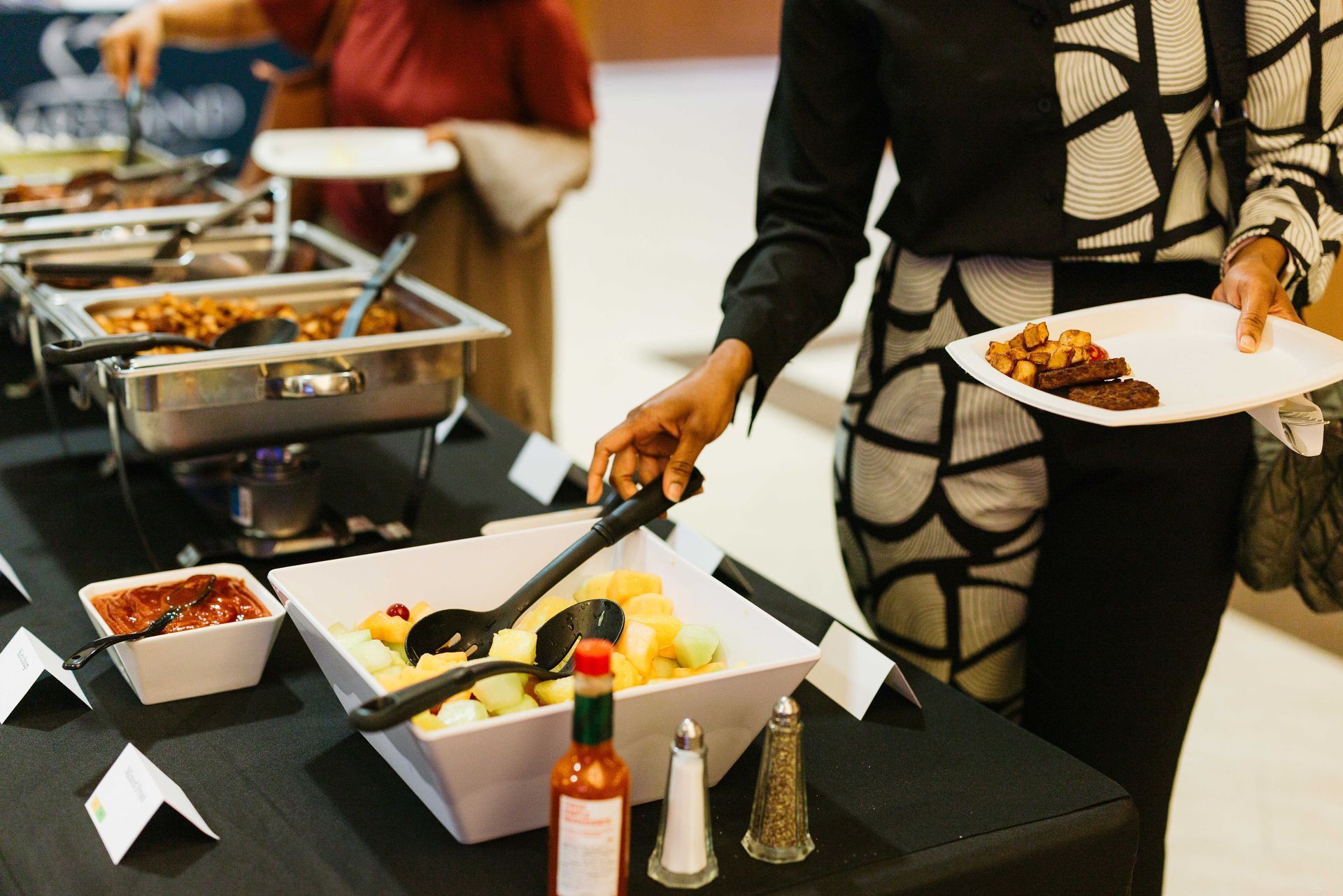 People serving food at a buffet table with tongs and plates in a dining area