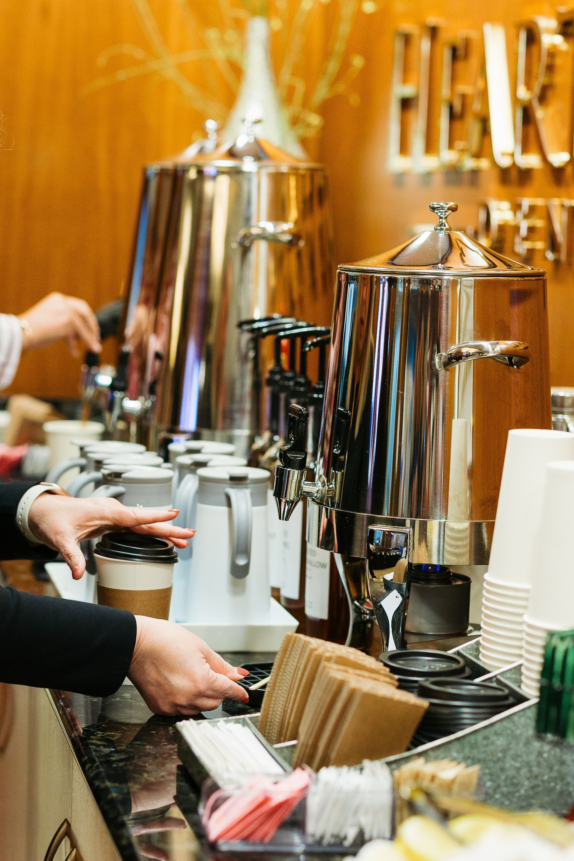 Coffee and tea service with chrome dispensers, cups, and hands pouring drinks at a buffet counter