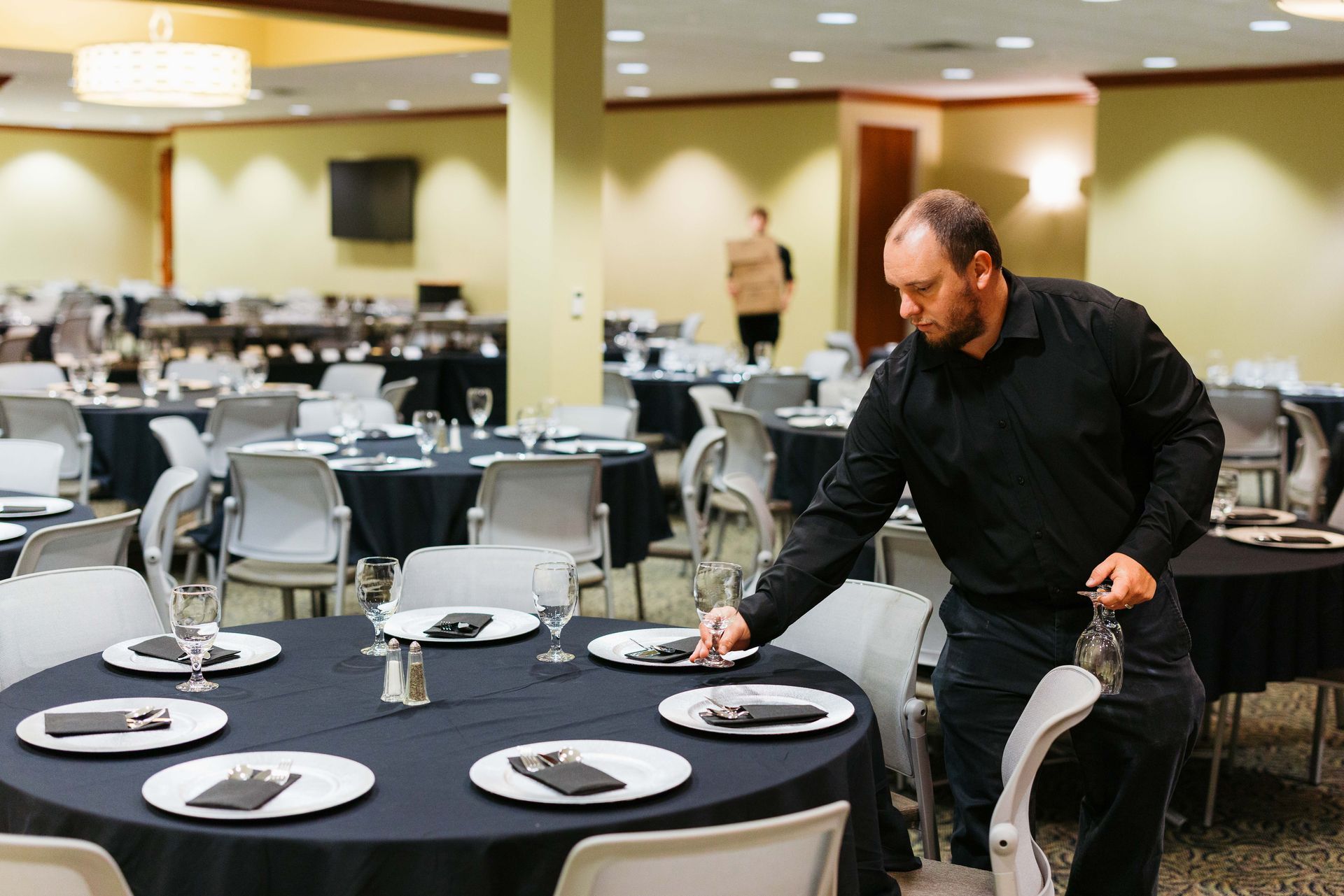 Man setting black tablecloths and place settings in a banquet hall with round tables and chairs