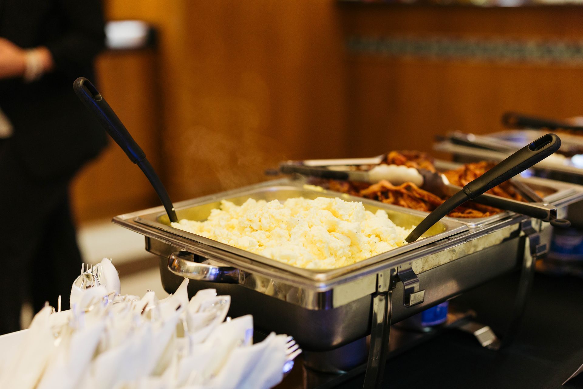 Buffet table with chafing dishes of food, including mashed potatoes and a meat dish, with serving utensils.