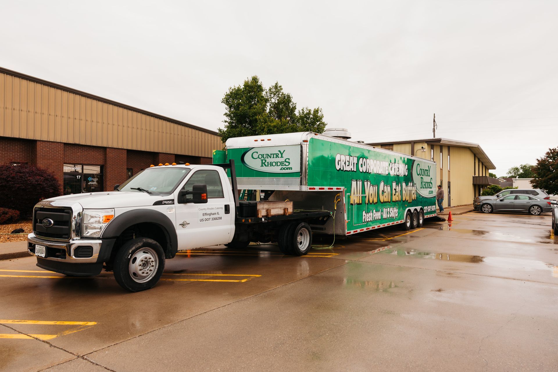 White truck with green dumpster trailer parked on a wet street outside a building