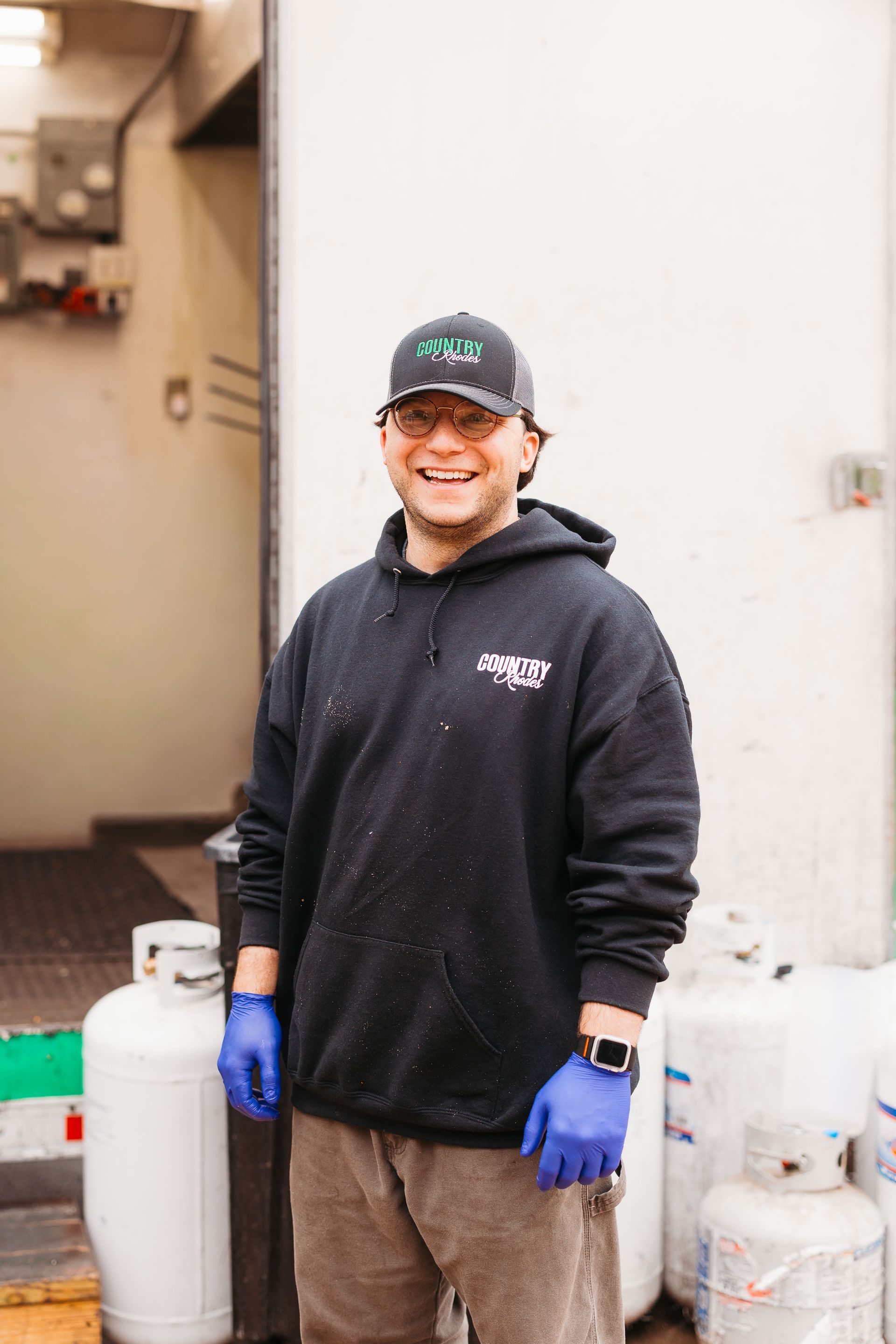 Smiling worker in black hoodie and blue gloves standing beside stacked white containers outside a building