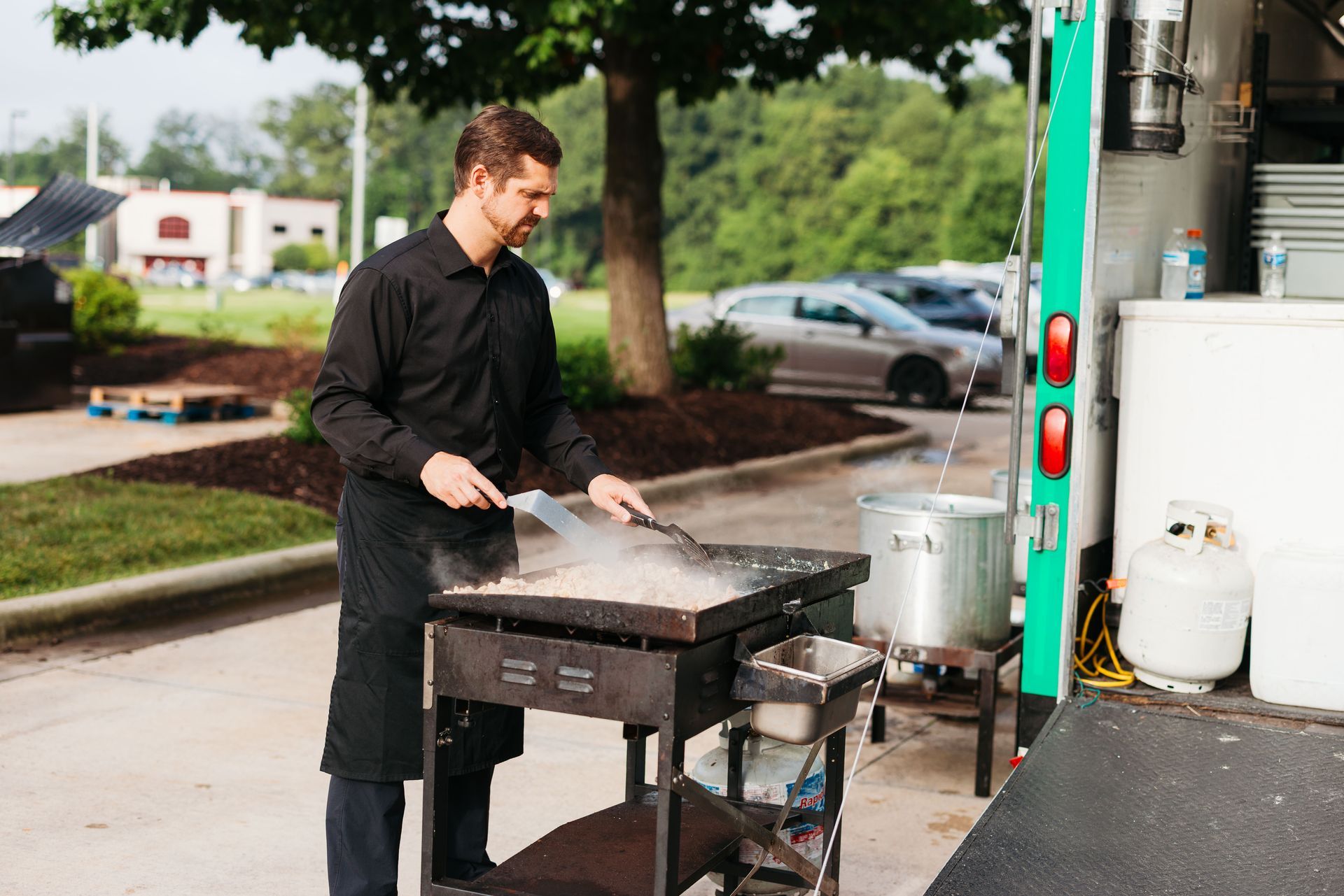 Person in a black coat cooking at a large outdoor grill beside a food truck.