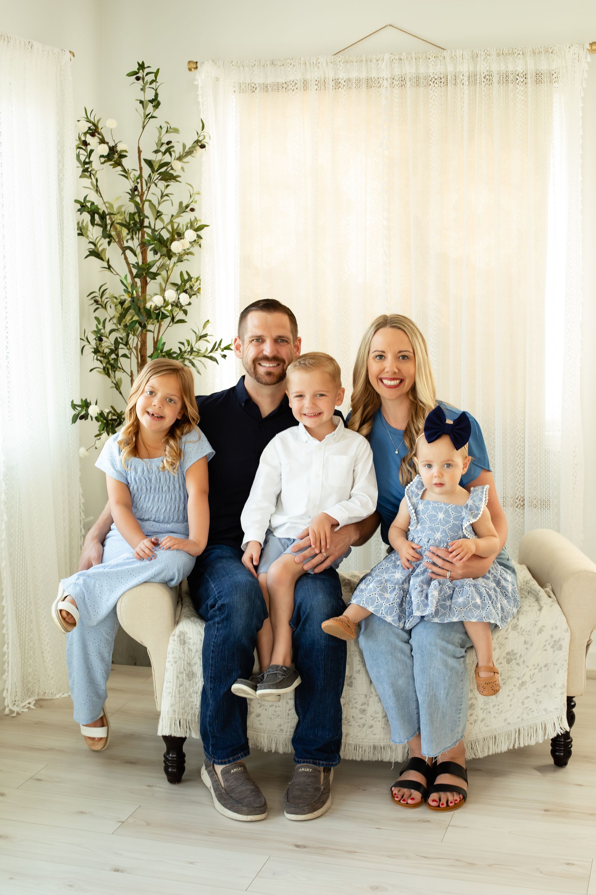 Family of five sitting on a cream sofa in a bright, white room with soft curtains and a tree branch backdrop.