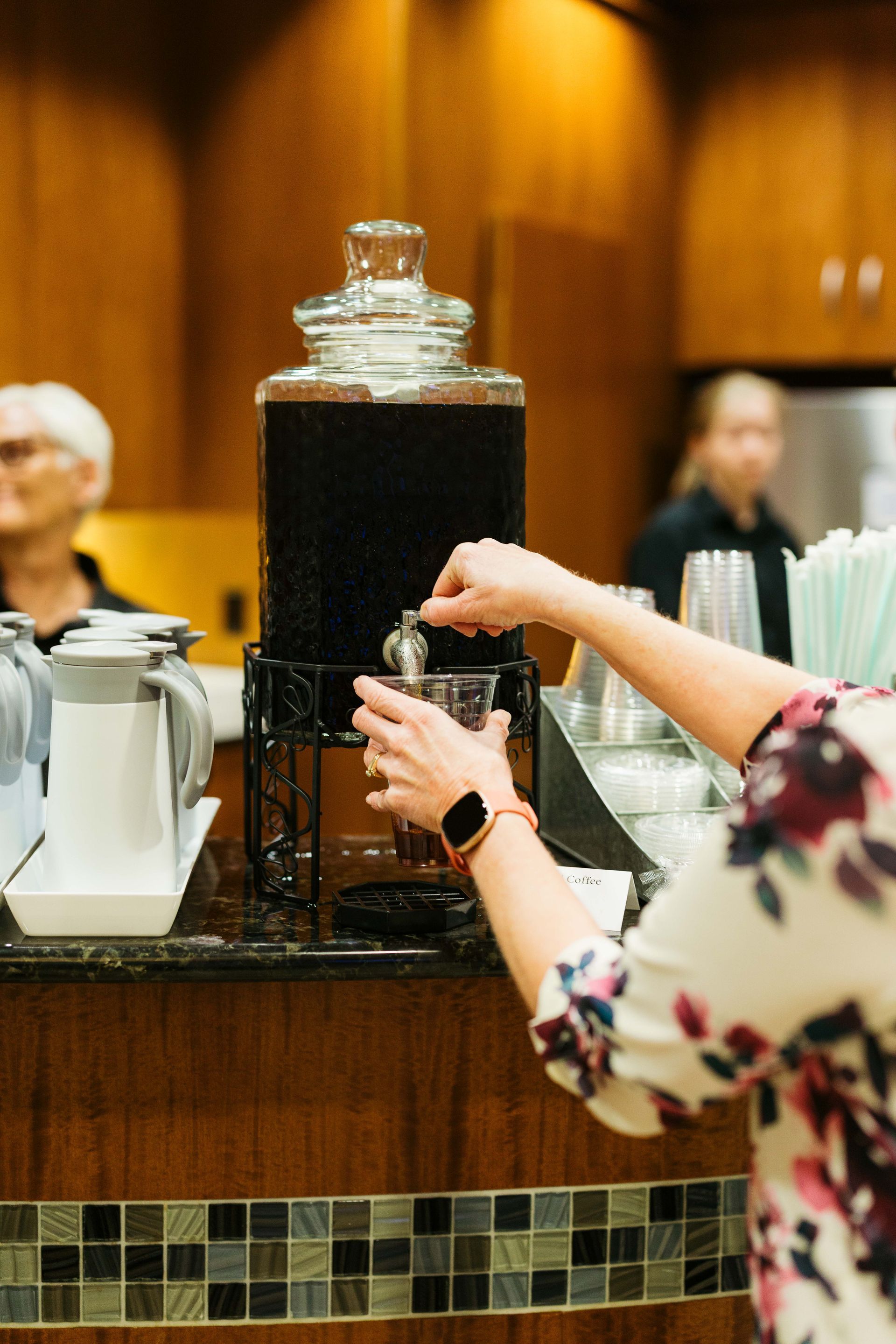 Hand placing a cup under a beverage dispenser in a warm-lit café or buffet area