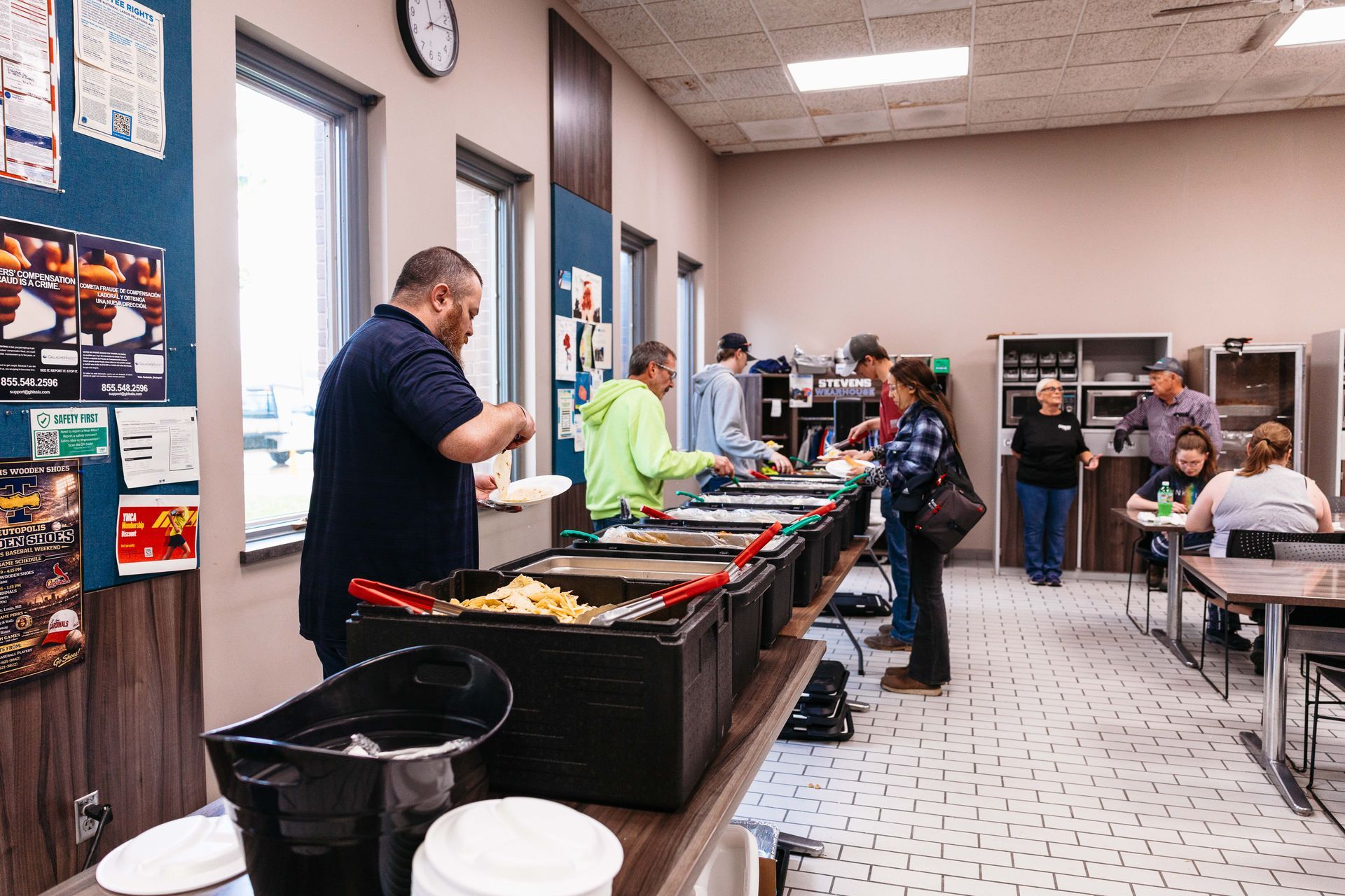 People serving food at a cafeteria buffet with diners seated at tables