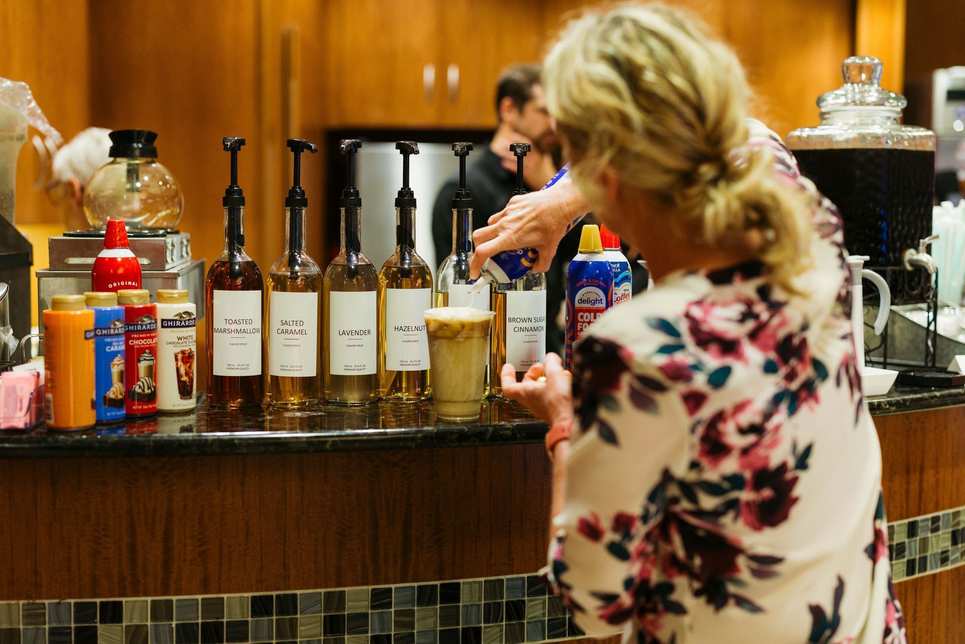 Bar counter with coffee dispensers and syrups; a blonde woman in a floral top stands at the counter.