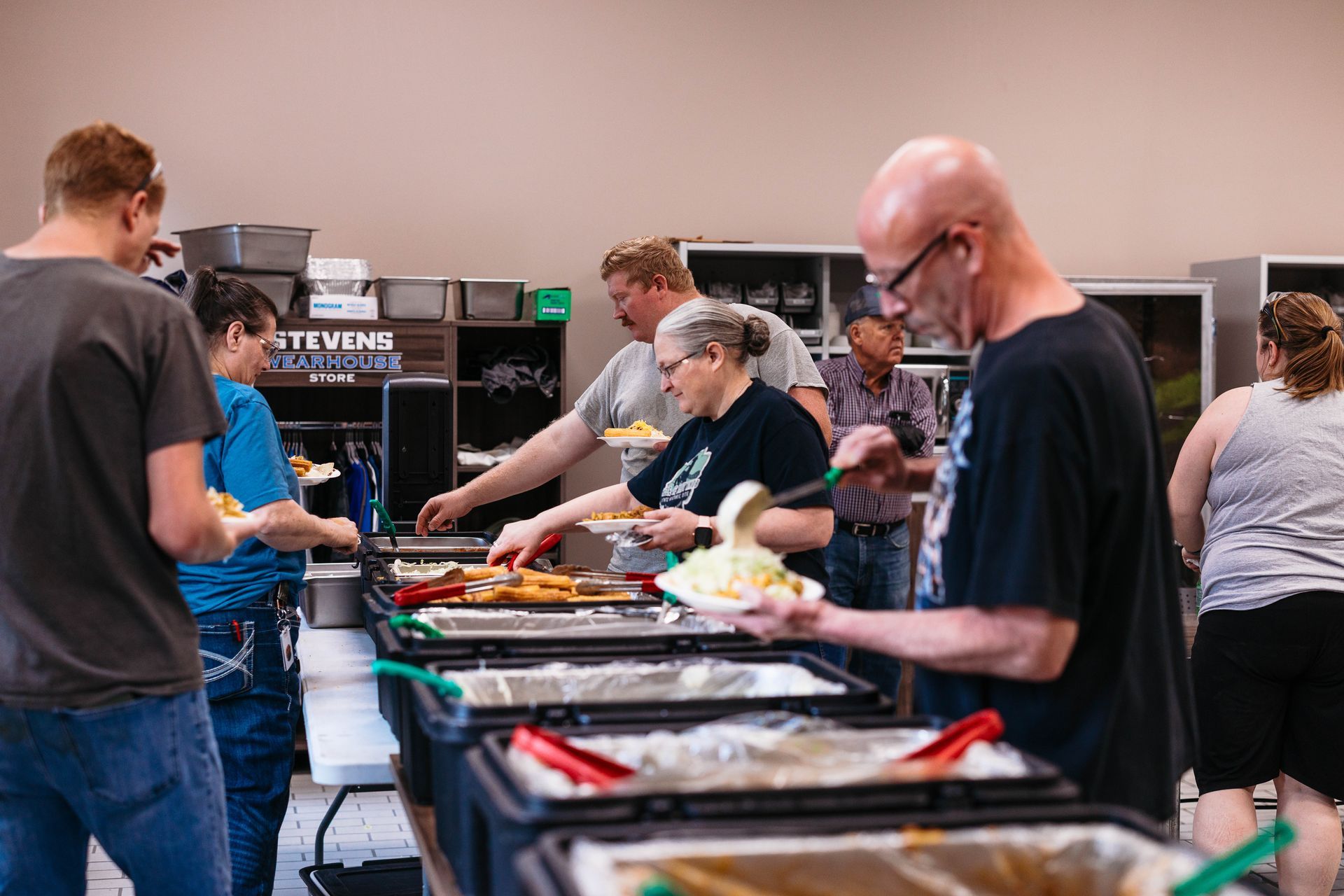 People serving themselves at a buffet table with trays of food in a cafeteria