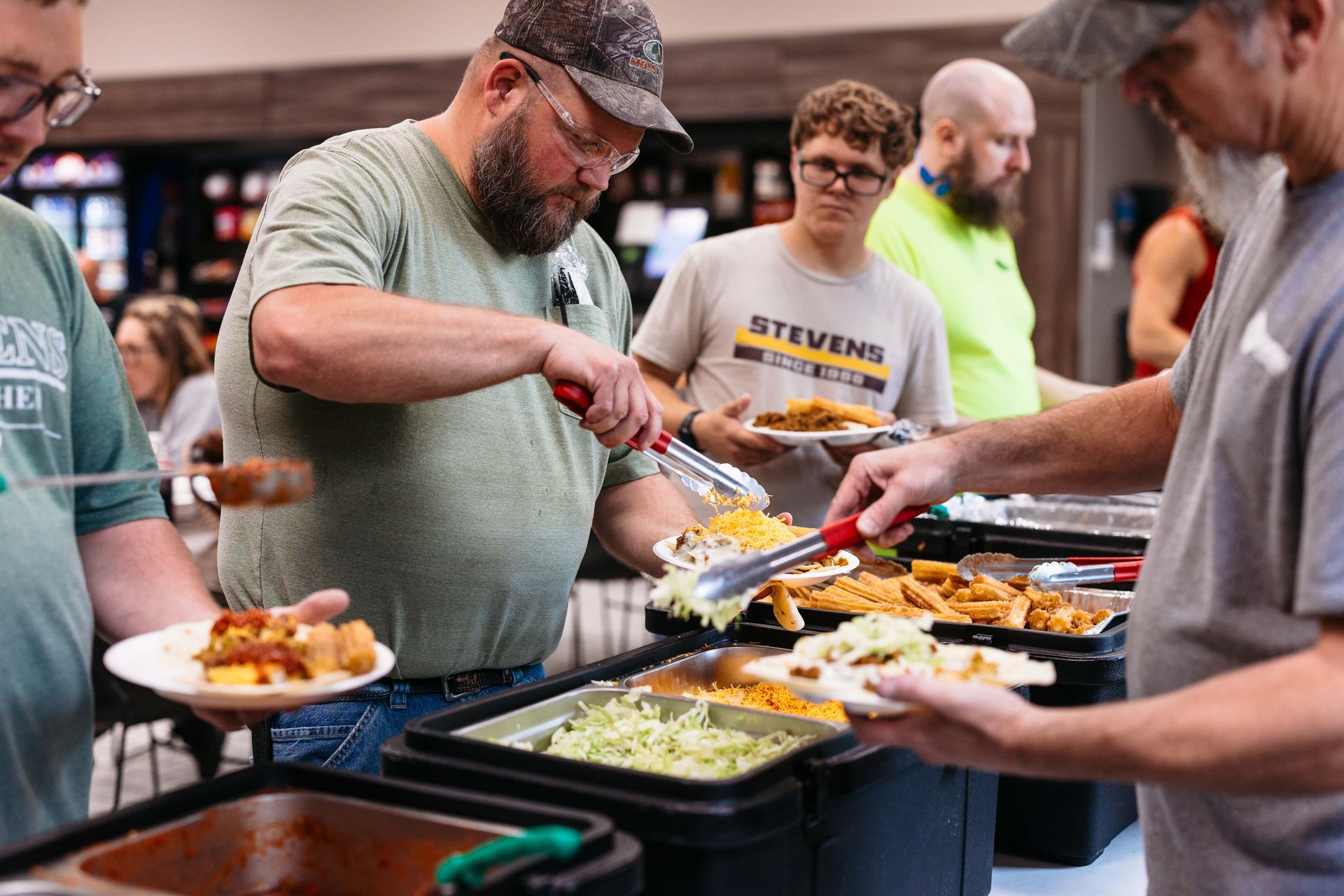 People serving themselves at a buffet table with trays of food in a cafeteria.