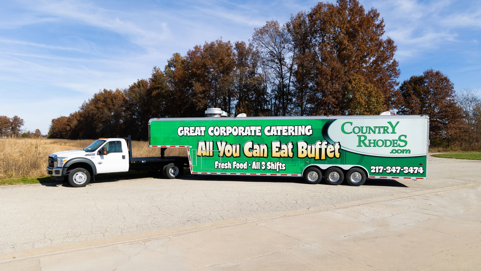White truck towing green Country Kitchen “All You Can Eat Buffet” trailer on a rural road.