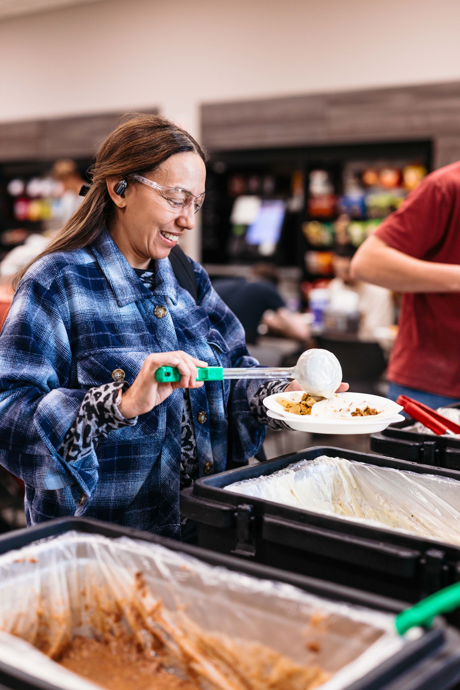 Person serving food from a buffet line with tongs, wearing a blue patterned jacket.