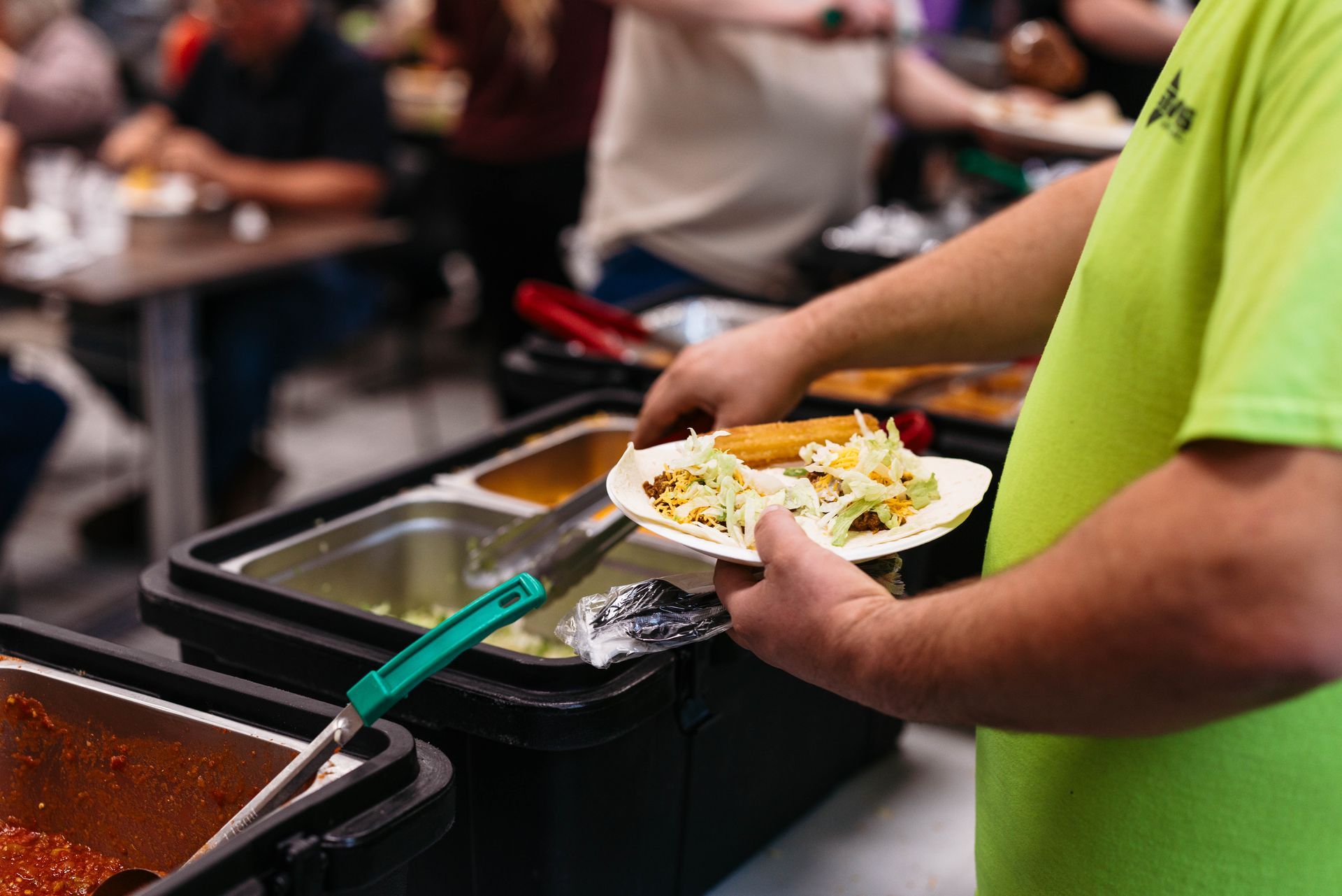Person serving food onto a plate from a buffet tray, wearing a bright green shirt.