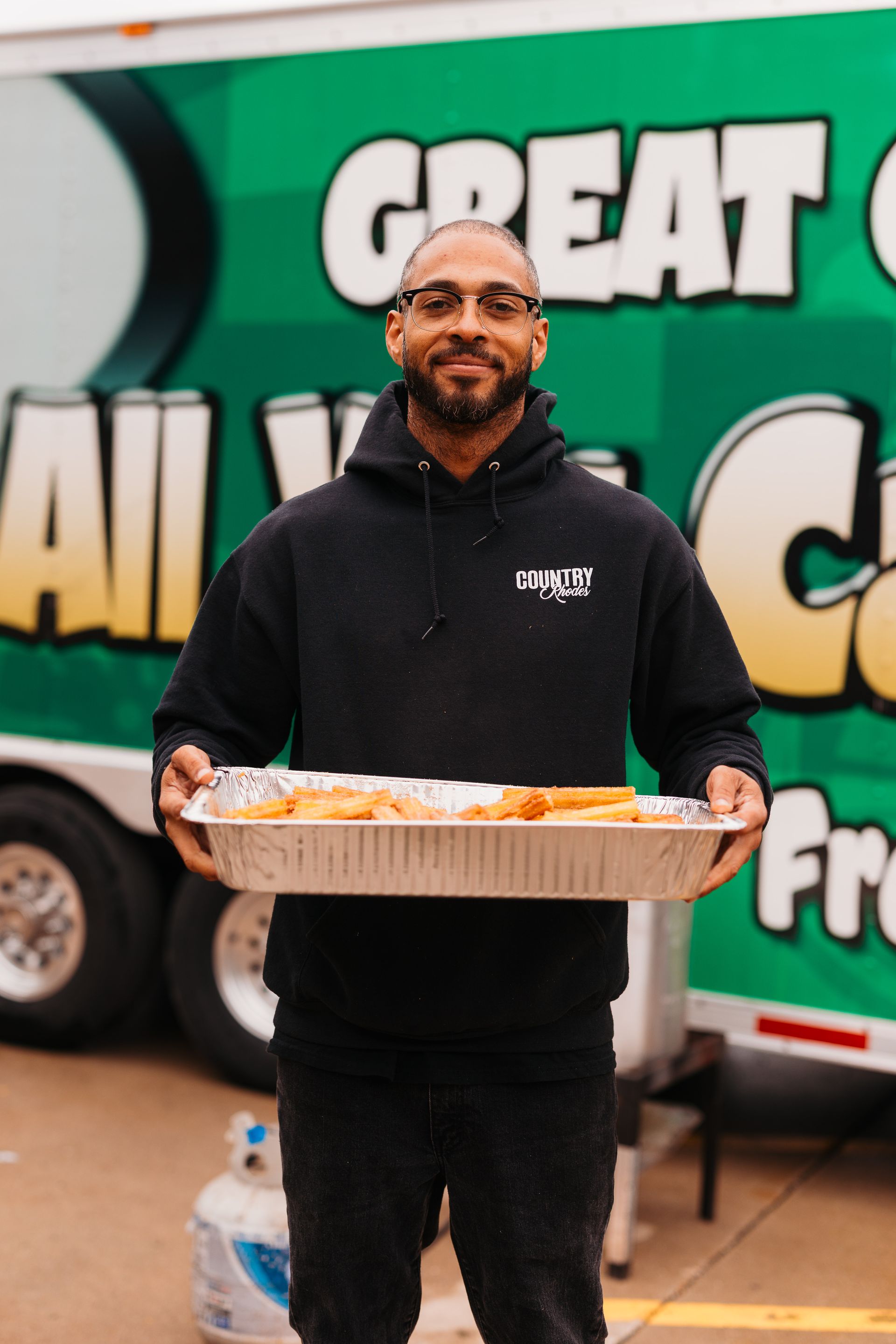 Man holding a tray of food in front of a green food truck with bold white lettering