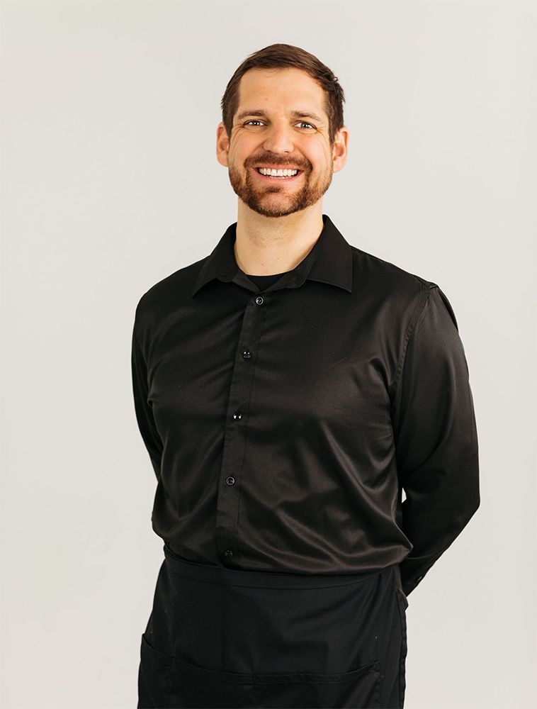 Smiling man in a black shirt standing against a light background, hands behind his back.