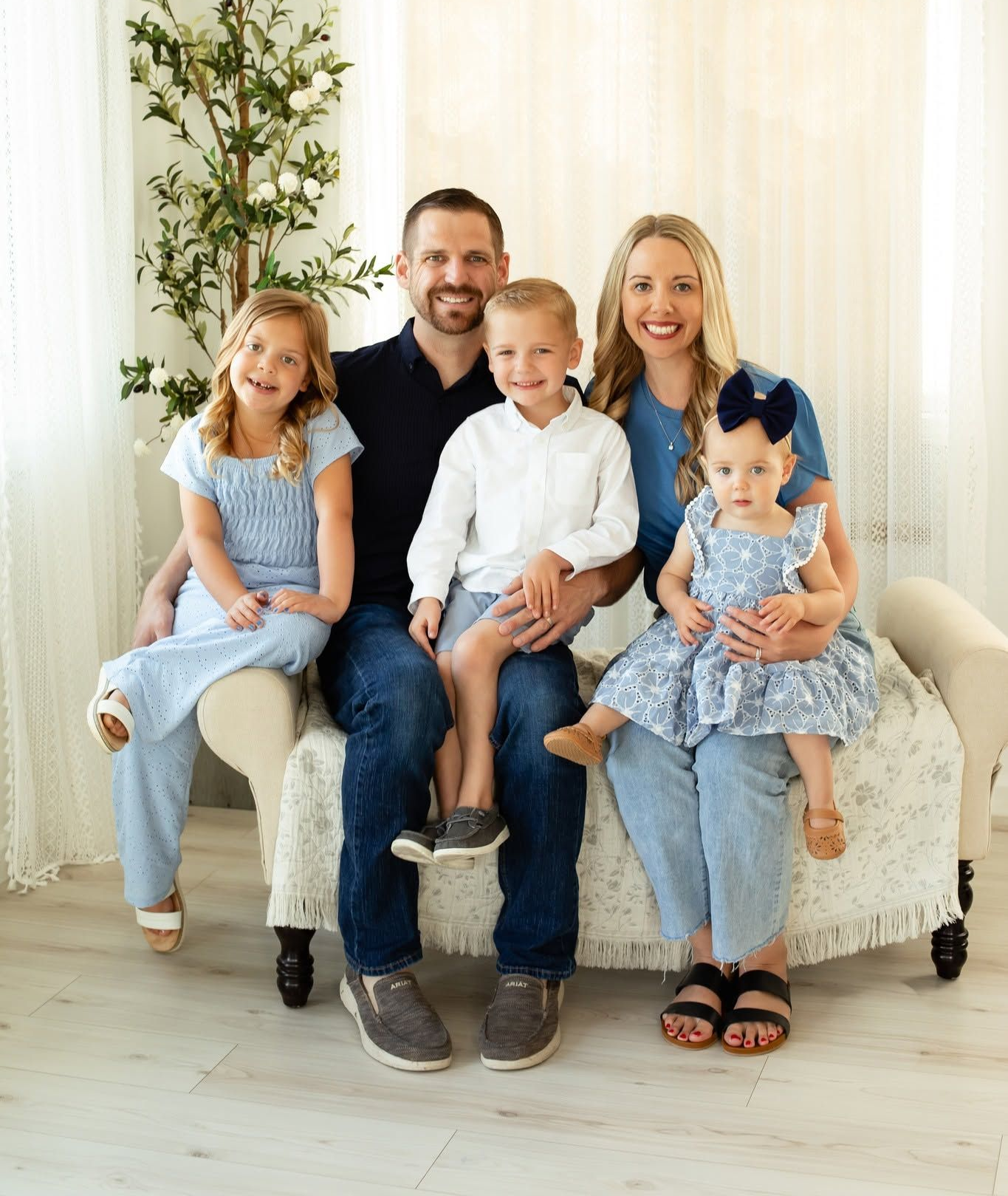Family of four sitting on a beige couch in a bright living room, smiling at the camera.