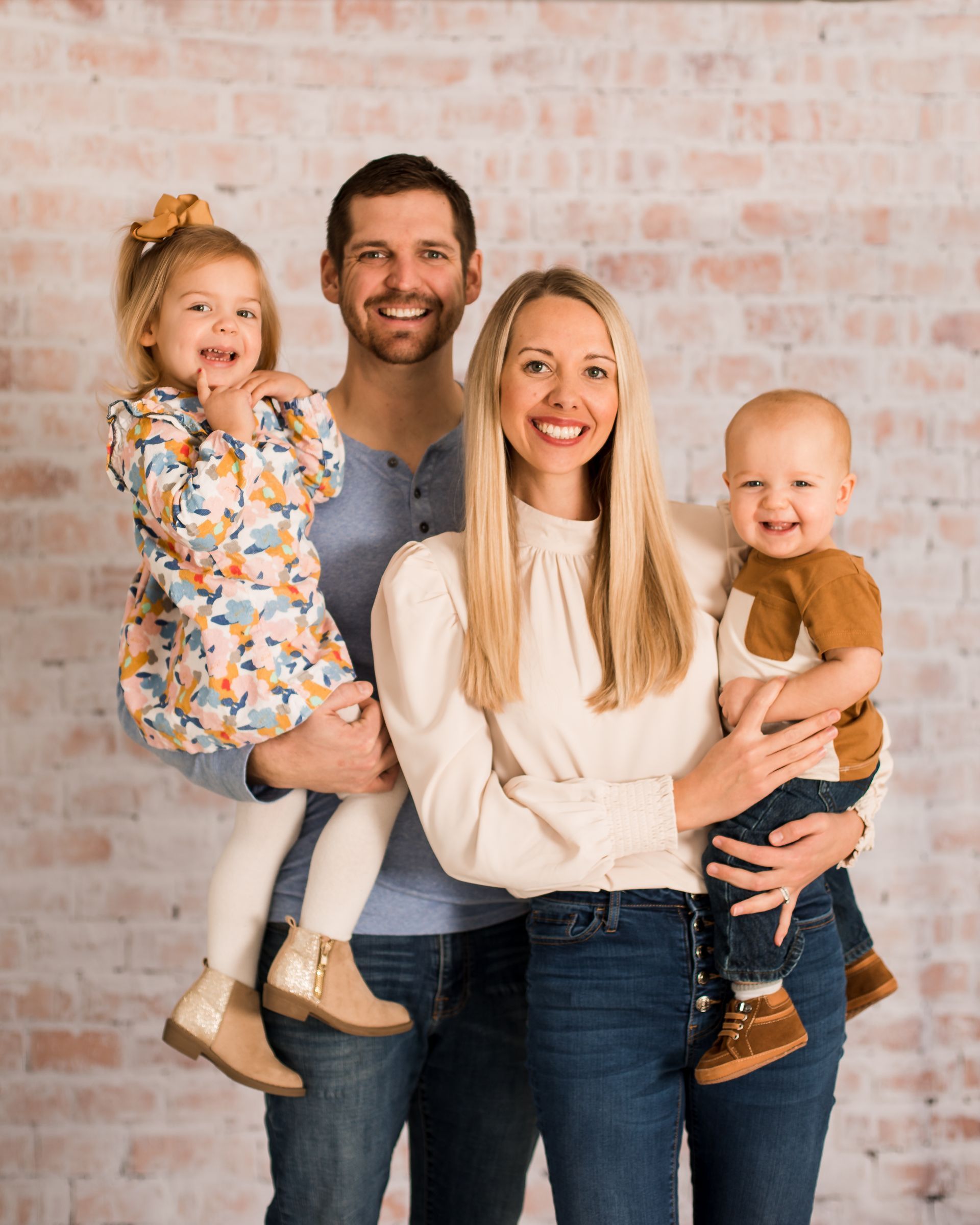 Family of four posing indoors against a light brick wall, smiling and holding two young children.