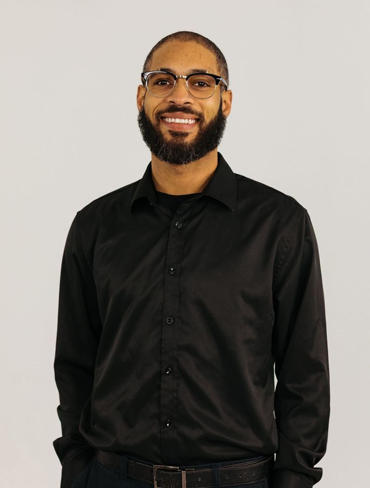 Smiling man in black shirt and glasses standing against a light background
