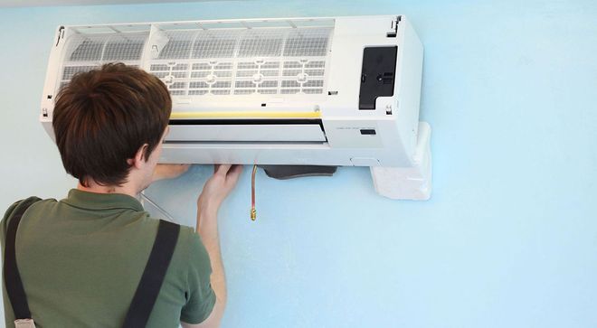 Person servicing a wall-mounted air conditioner with its cover open and tools hanging below.