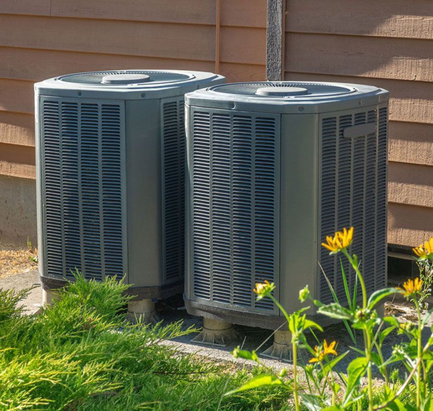 Two outdoor central air conditioner units beside a house wall, with grass and yellow flowers in front