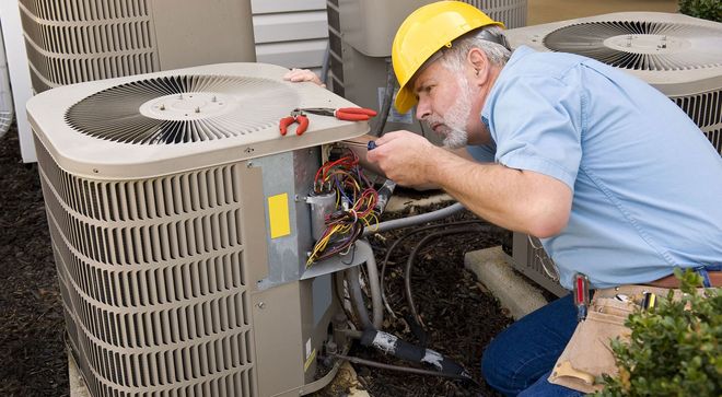Technician in yellow hard hat repairing outdoor air conditioner unit with exposed wiring