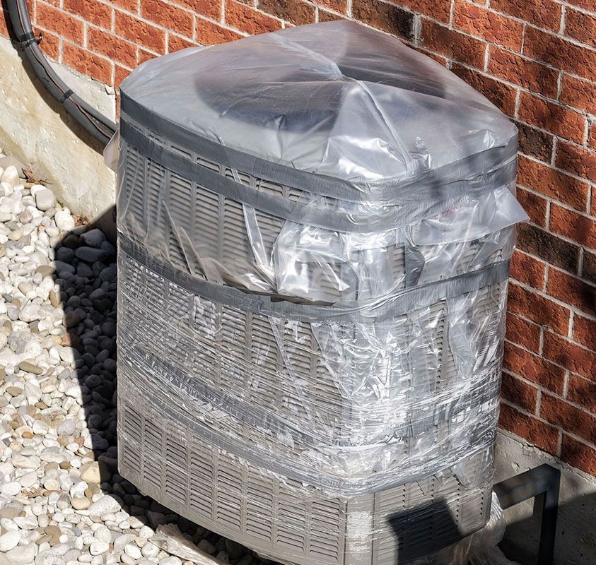 Outdoor HVAC unit wrapped in clear plastic beside a brick wall and white stones