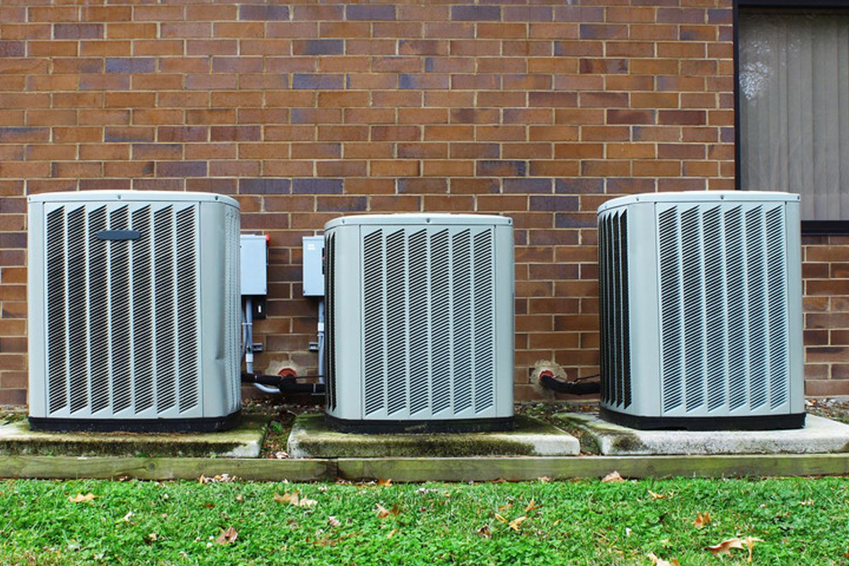Three outdoor air conditioning units beside a brick wall, with grass in front.