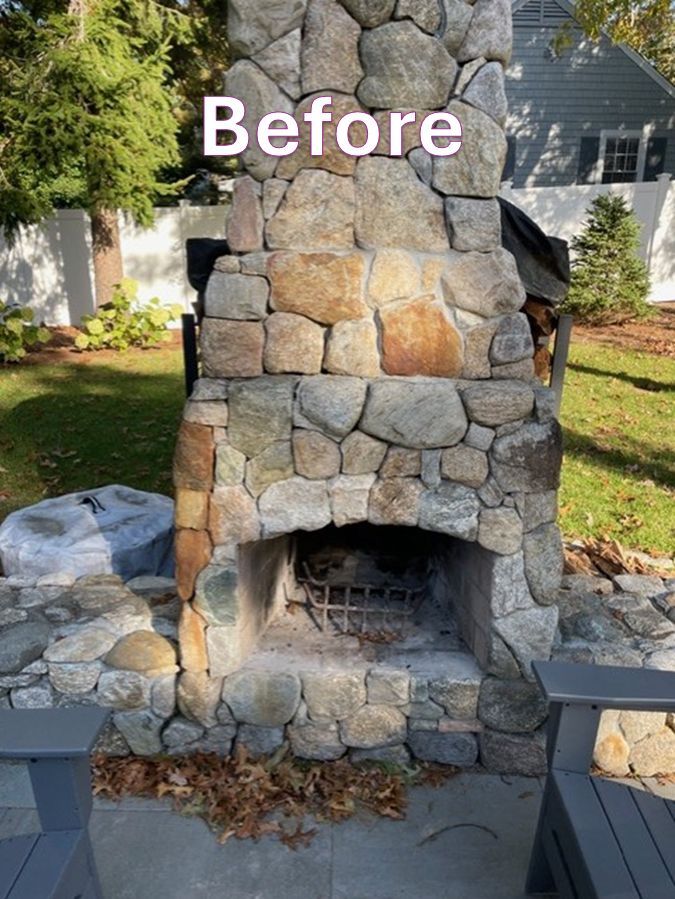 Stone outdoor fireplace; Light-colored stones, rectangular opening, surrounded by greenery.