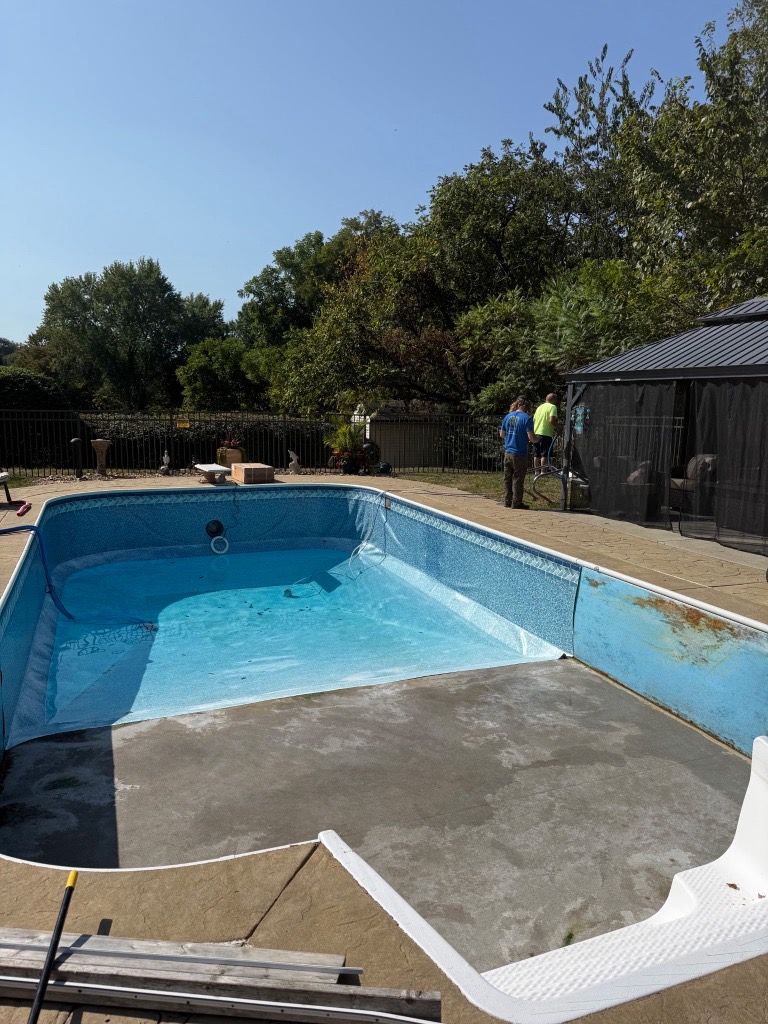 Empty backyard swimming pool being cleaned, with two workers near a shed on a sunny day