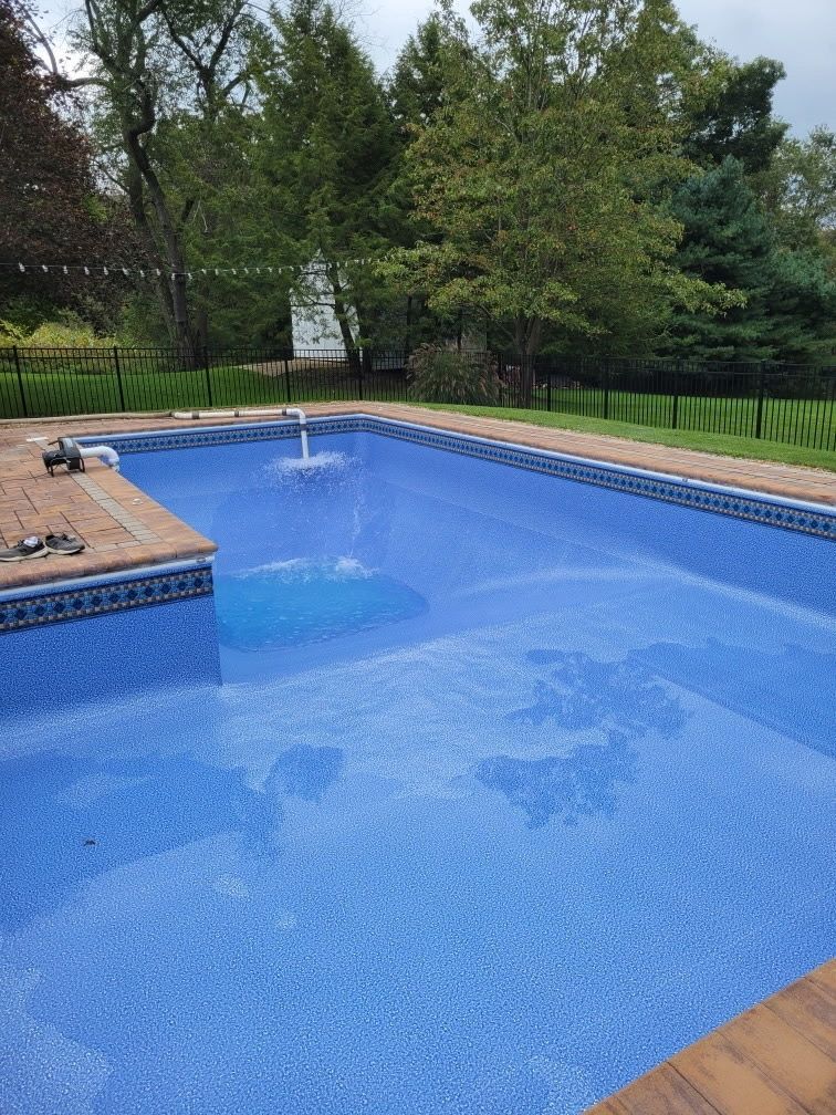 Blue backyard swimming pool with a fountain, tan deck, and green trees in the background.