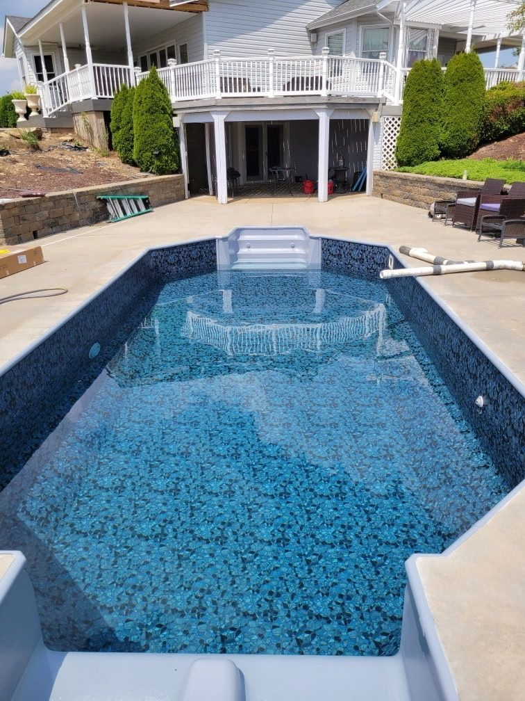 Backyard swimming pool with blue water, stone deck, and a house with white railings and patio seating.