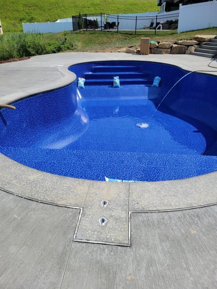 Curved blue in-ground pool with a concrete deck and spa steps beside a grassy backyard.