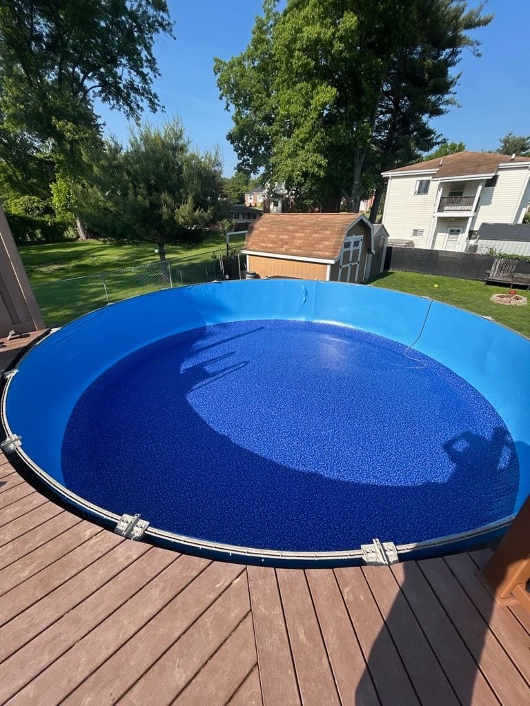 Blue above-ground pool on a wooden deck in a sunny backyard with trees and a house.