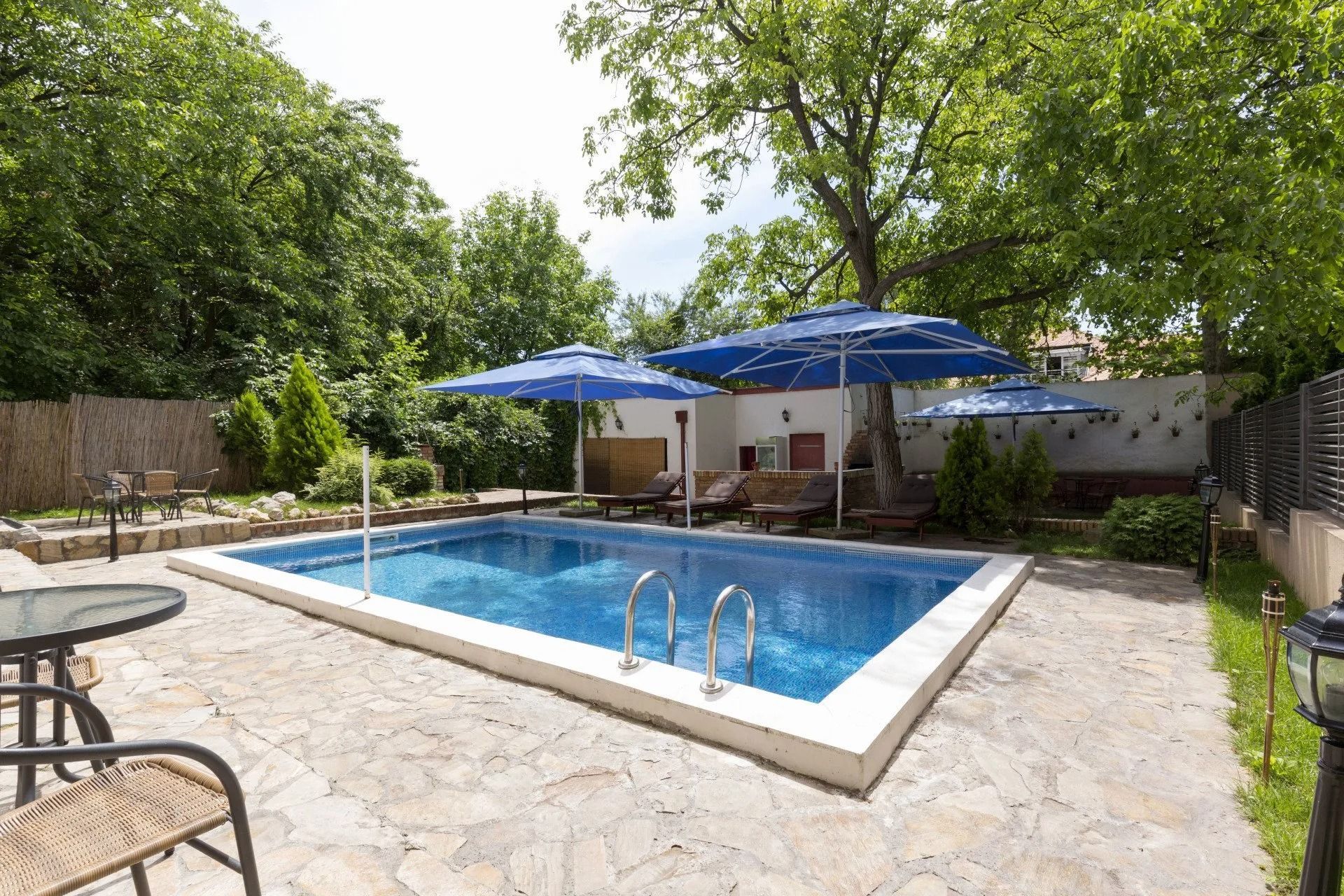 Backyard swimming pool with blue umbrellas, stone patio, and lush green trees