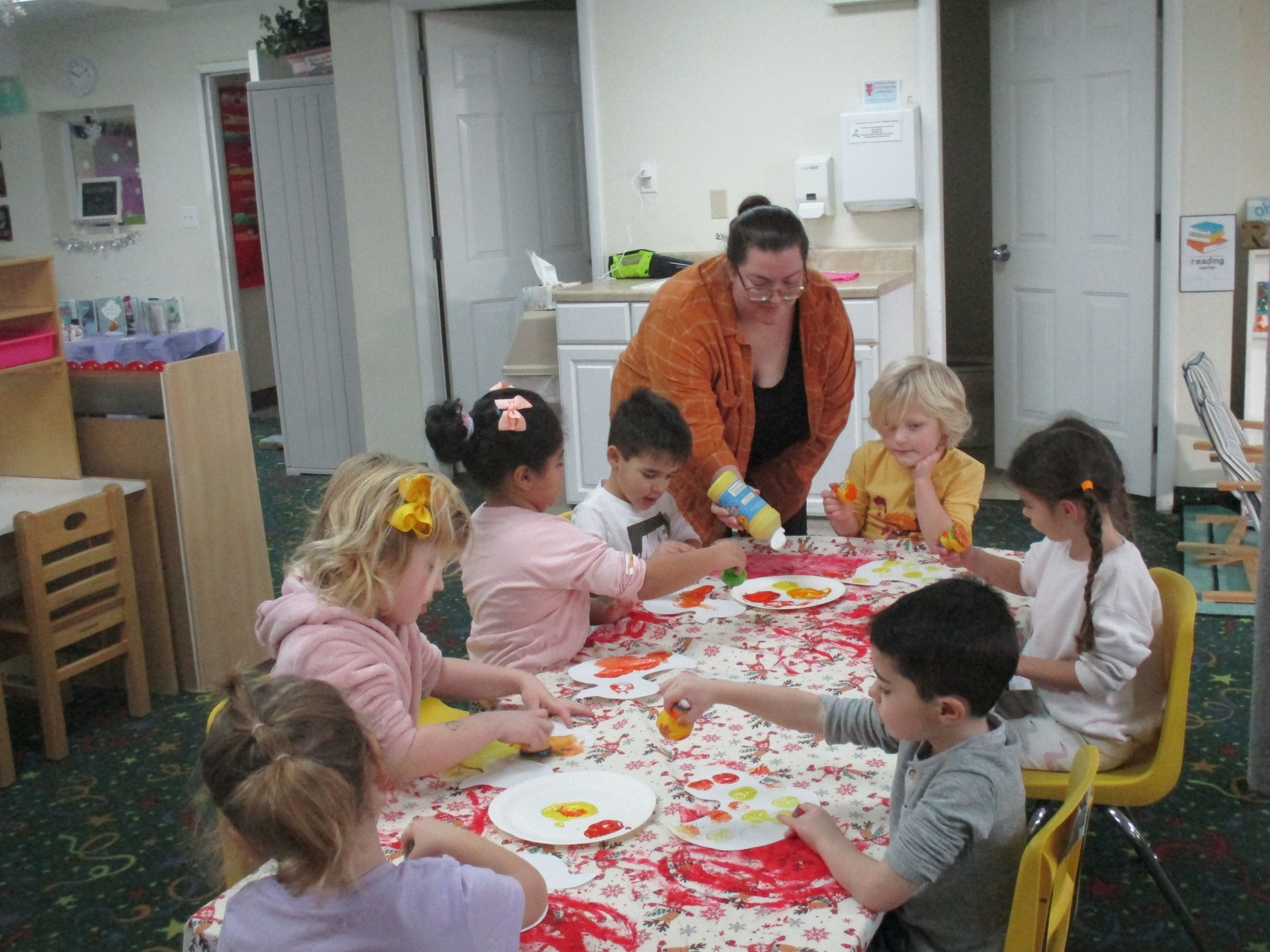 A group of children are sitting around a table with a woman standing behind them.