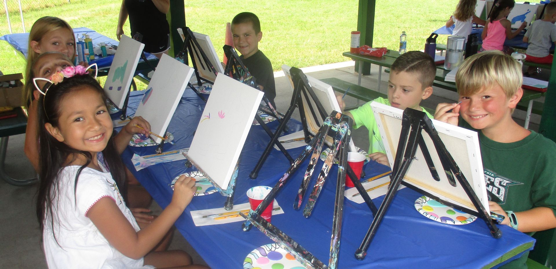 A group of children are sitting at a table painting on canvases.