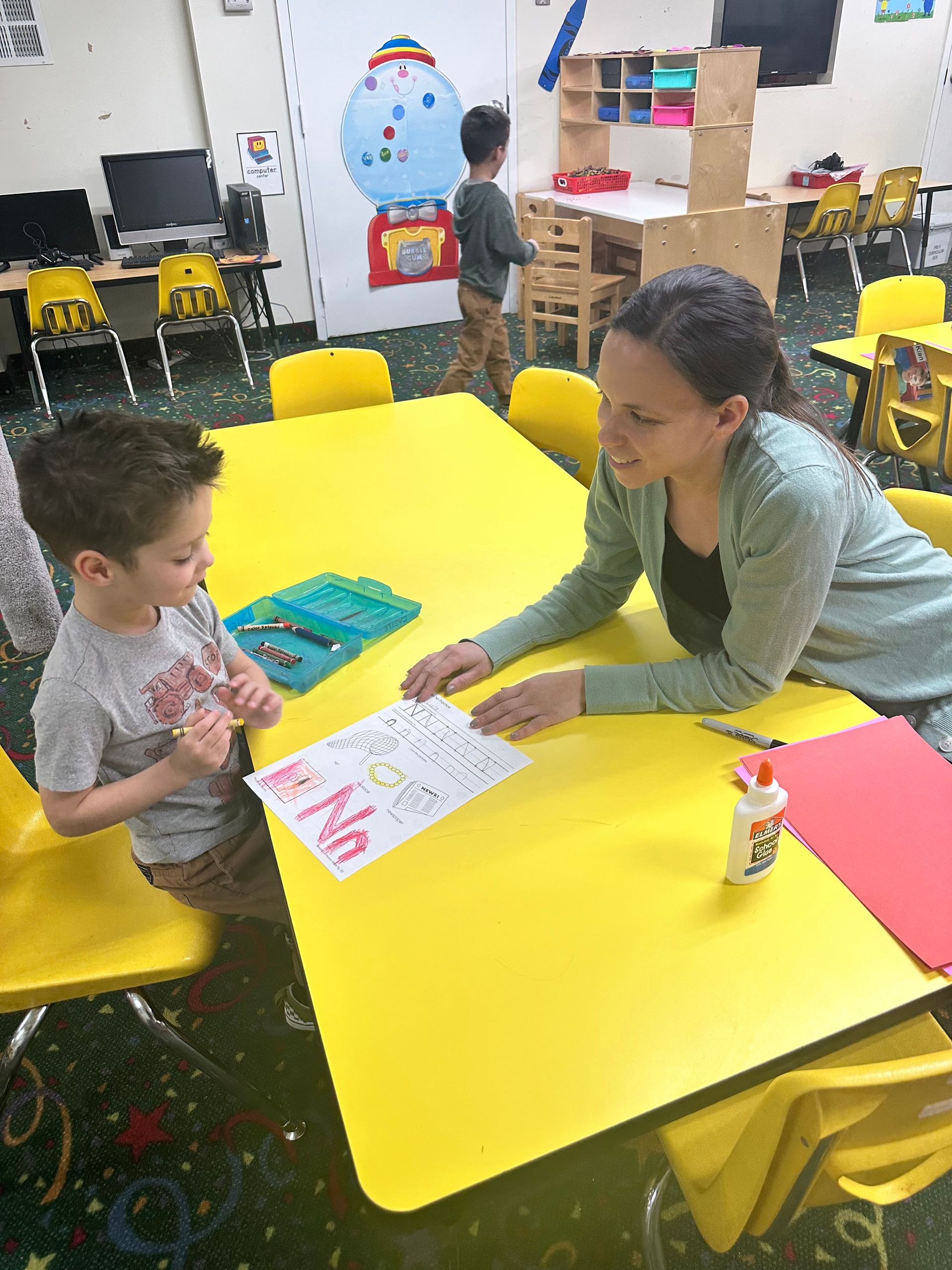 A woman is sitting at a table with a child in a classroom.