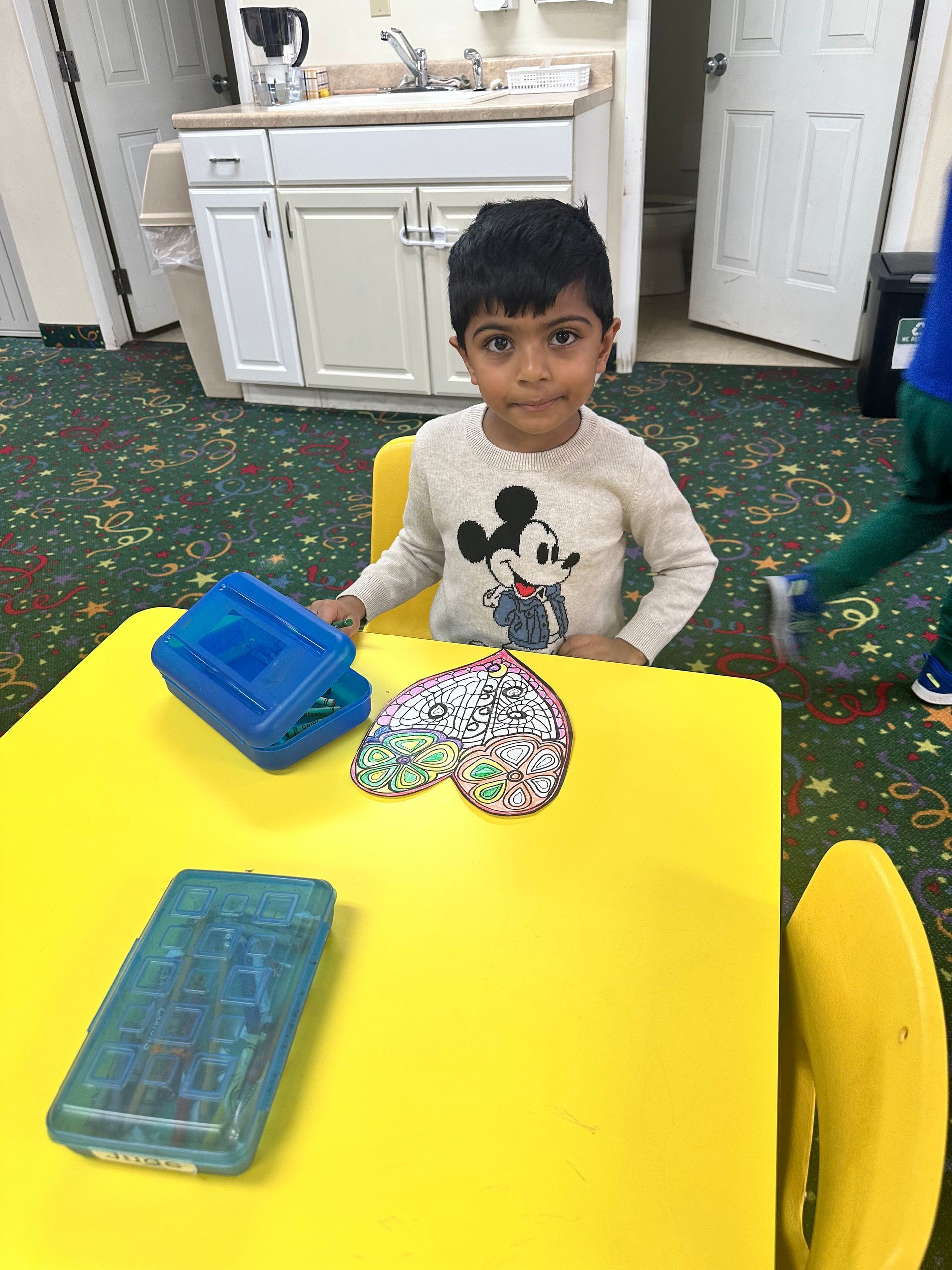 A young boy is sitting at a yellow table with a mickey mouse sweater on.