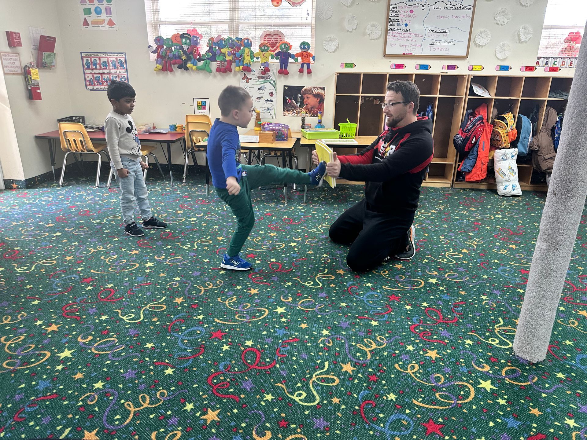 Teacher assisting a child kicking a held target in a preschool classroom.