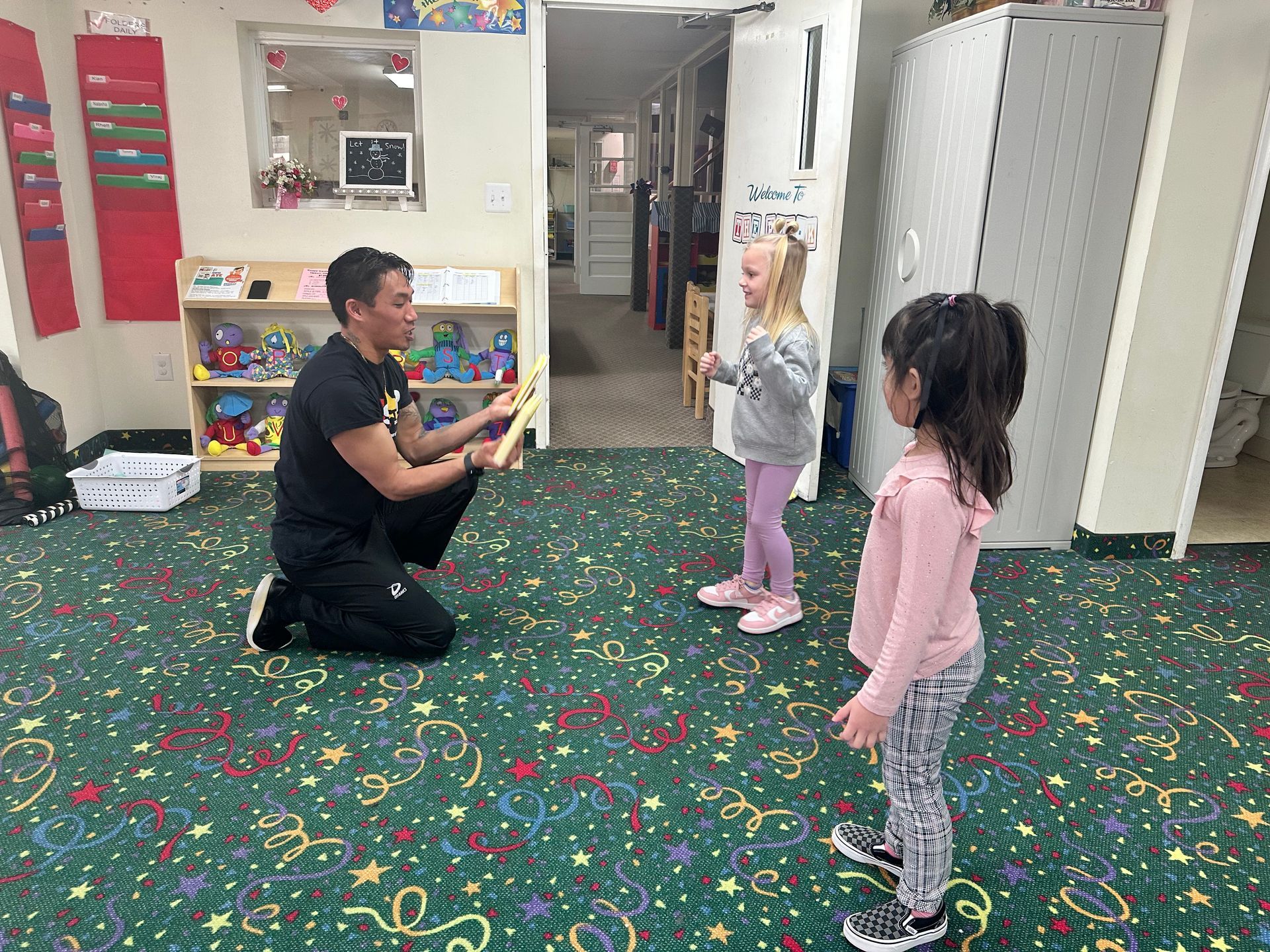 Man showing items to two young girls on a colorful carpet in a classroom.
