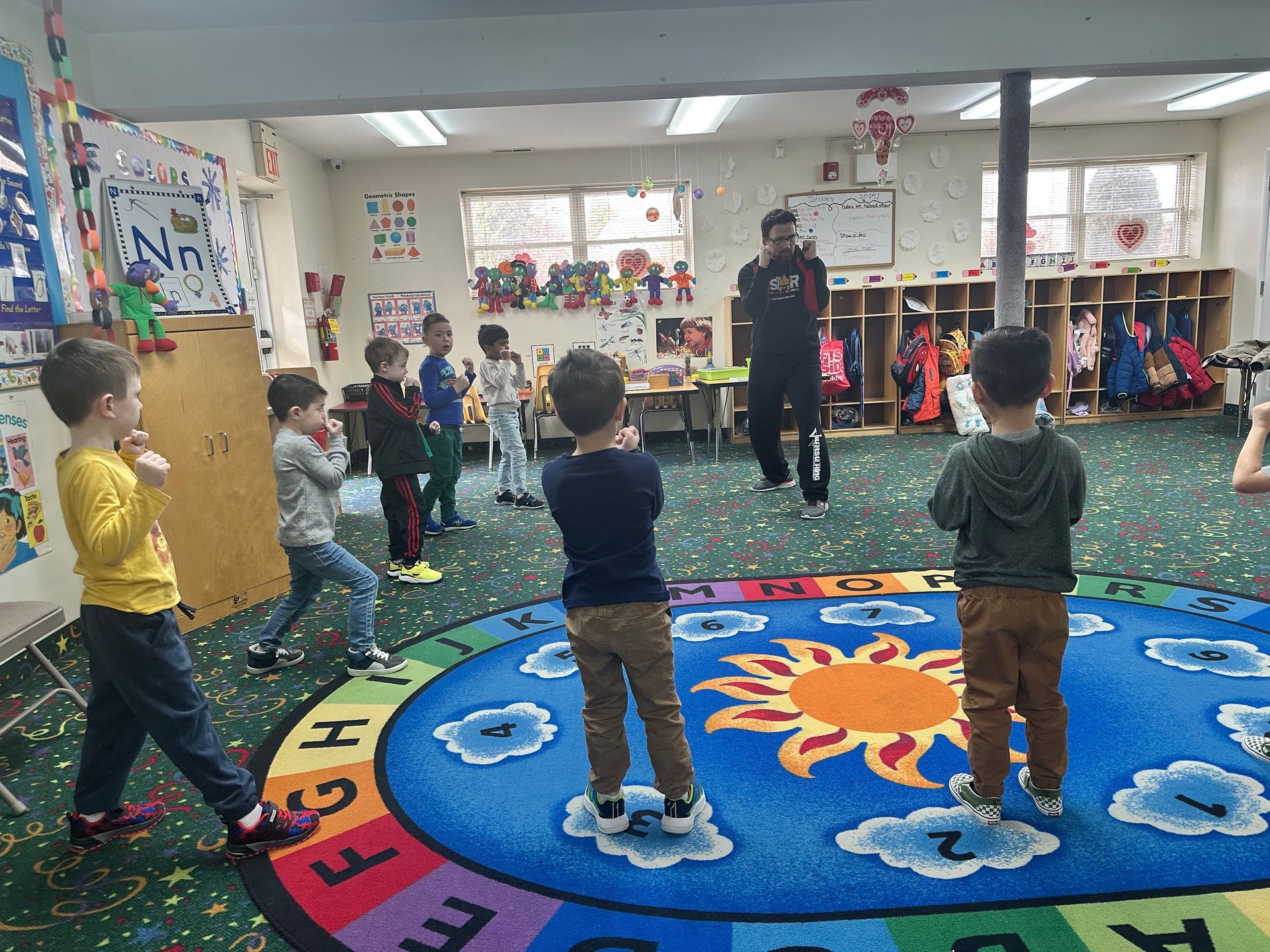 A group of children are standing in a circle on a rug in a classroom.