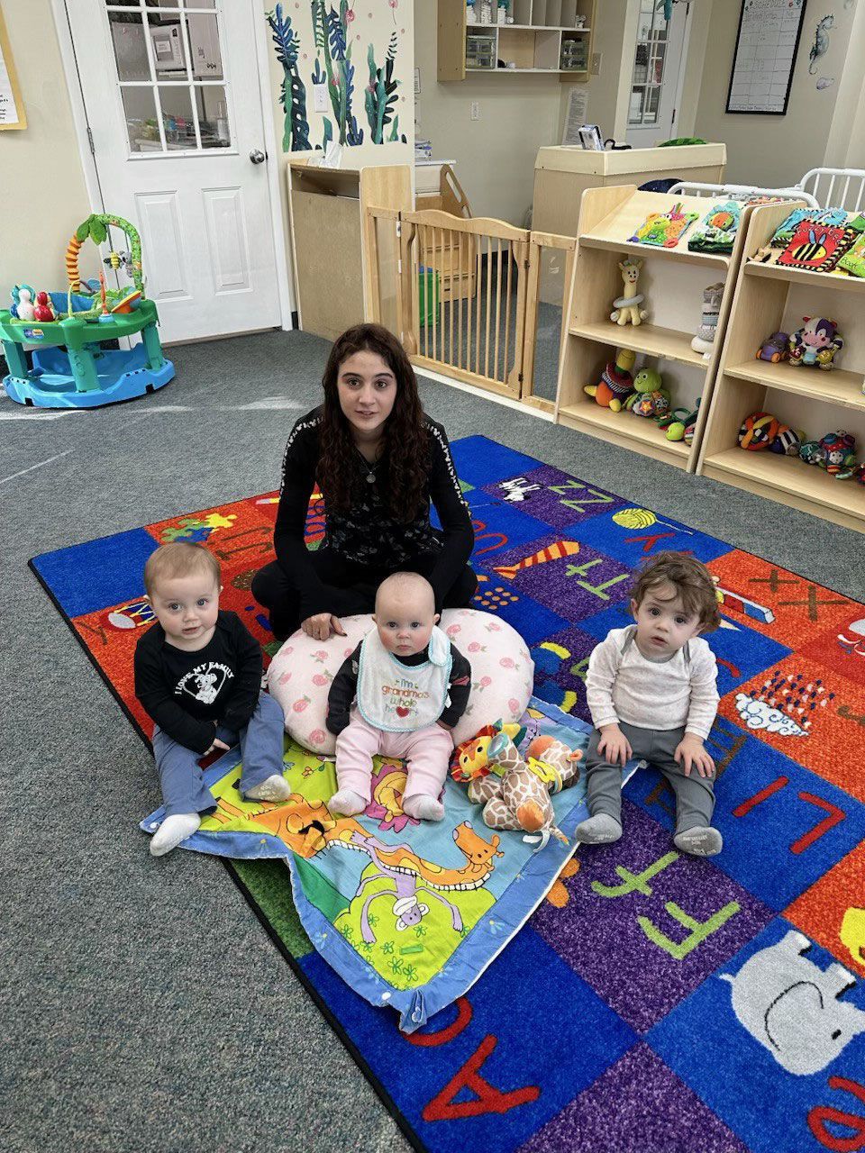 A woman sits with three babies on a colorful rug in a daycare