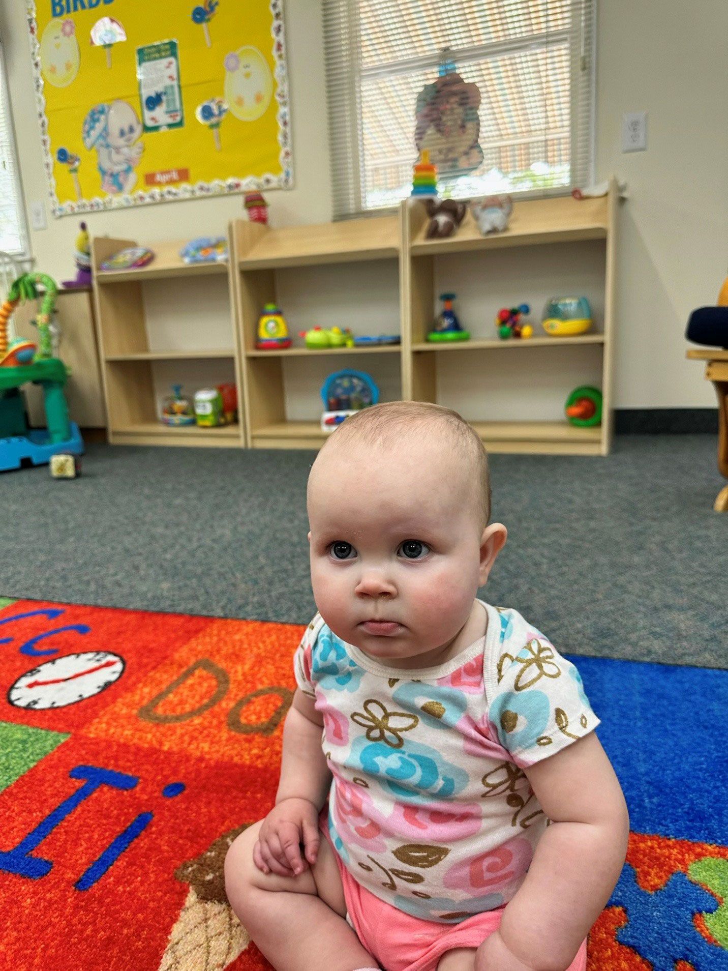 A baby sits on a colorful rug in a classroom, looking slightly grumpy