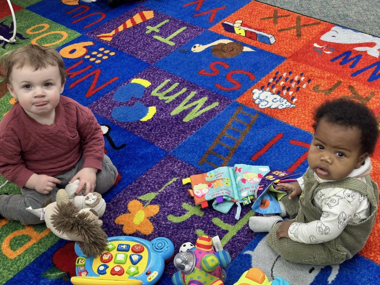 Two toddlers sit on an alphabet-themed rug with toys