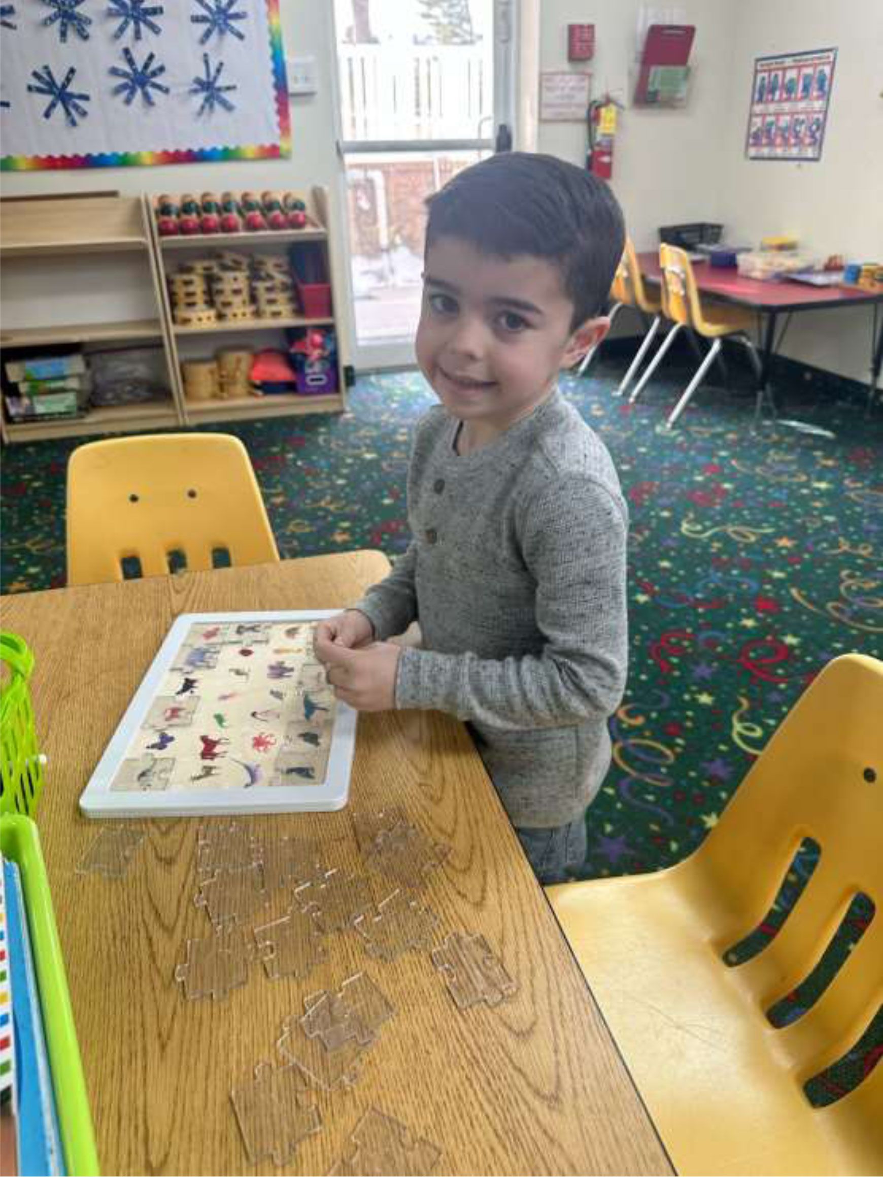 A young boy smiles at the camera, standing next to a wooden table in a classroom.