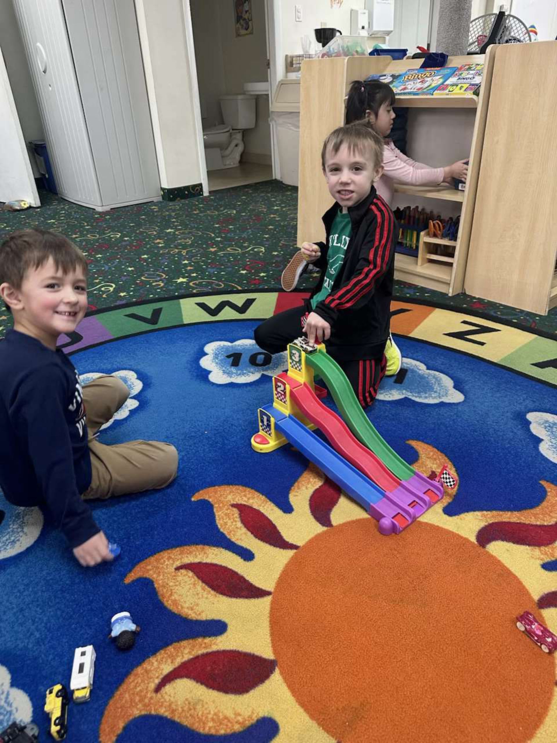 Two young boys play with a toy race track on a colorful rug.