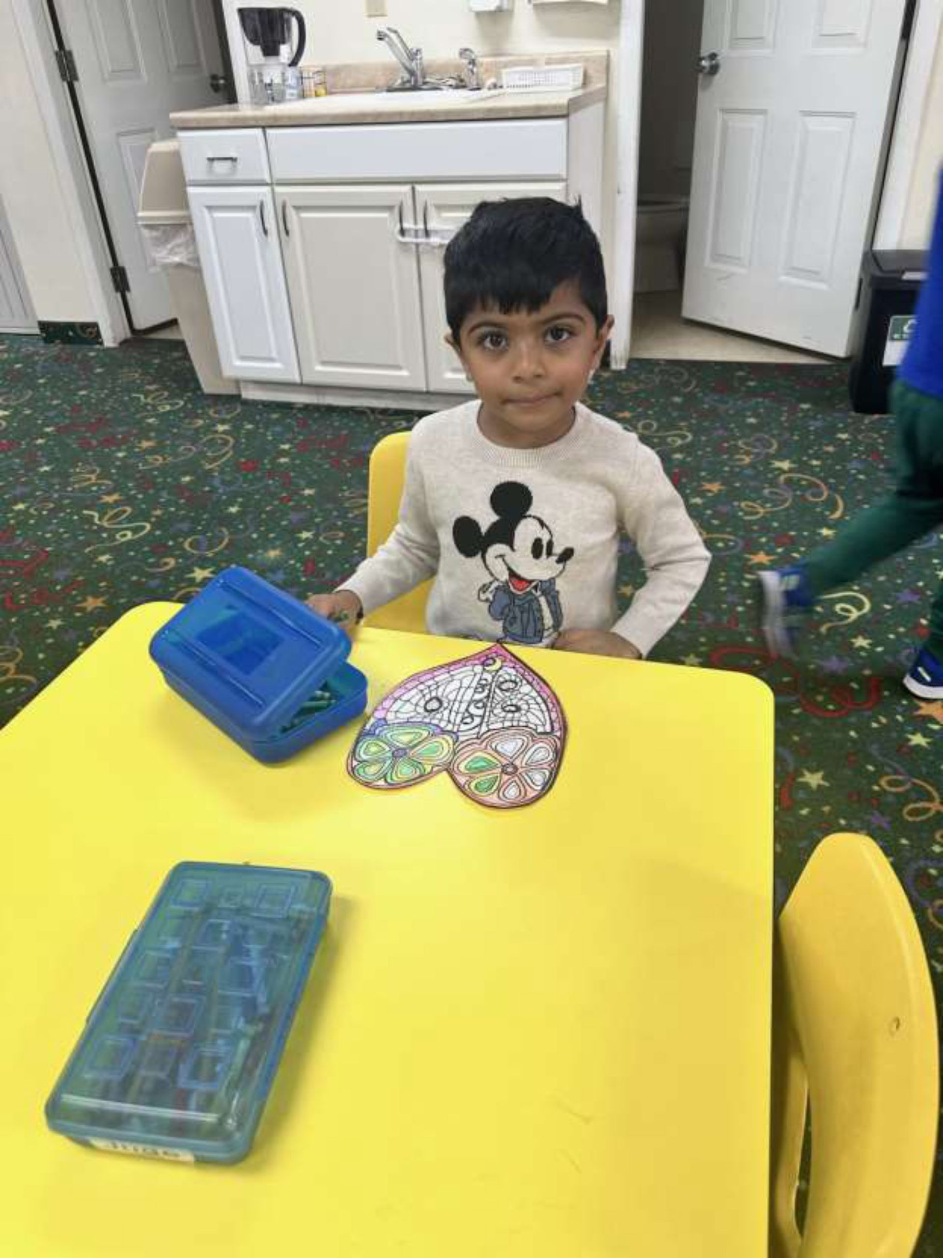 A young boy with dark hair and a Mickey Mouse sweater sits at a yellow table.