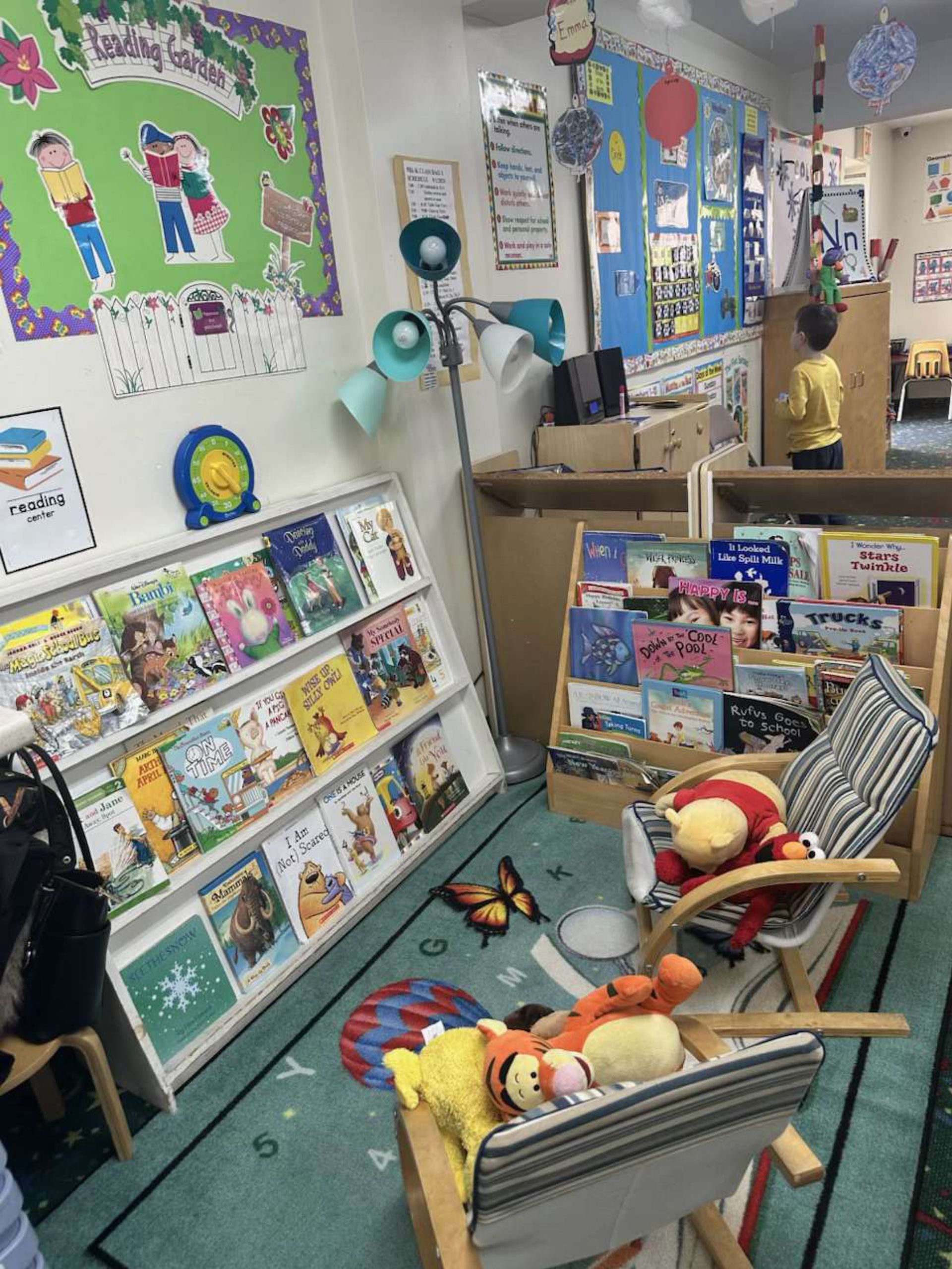 A child's reading corner: bookshelves with books, a rocking chair with plush toys, and a child in the background.
